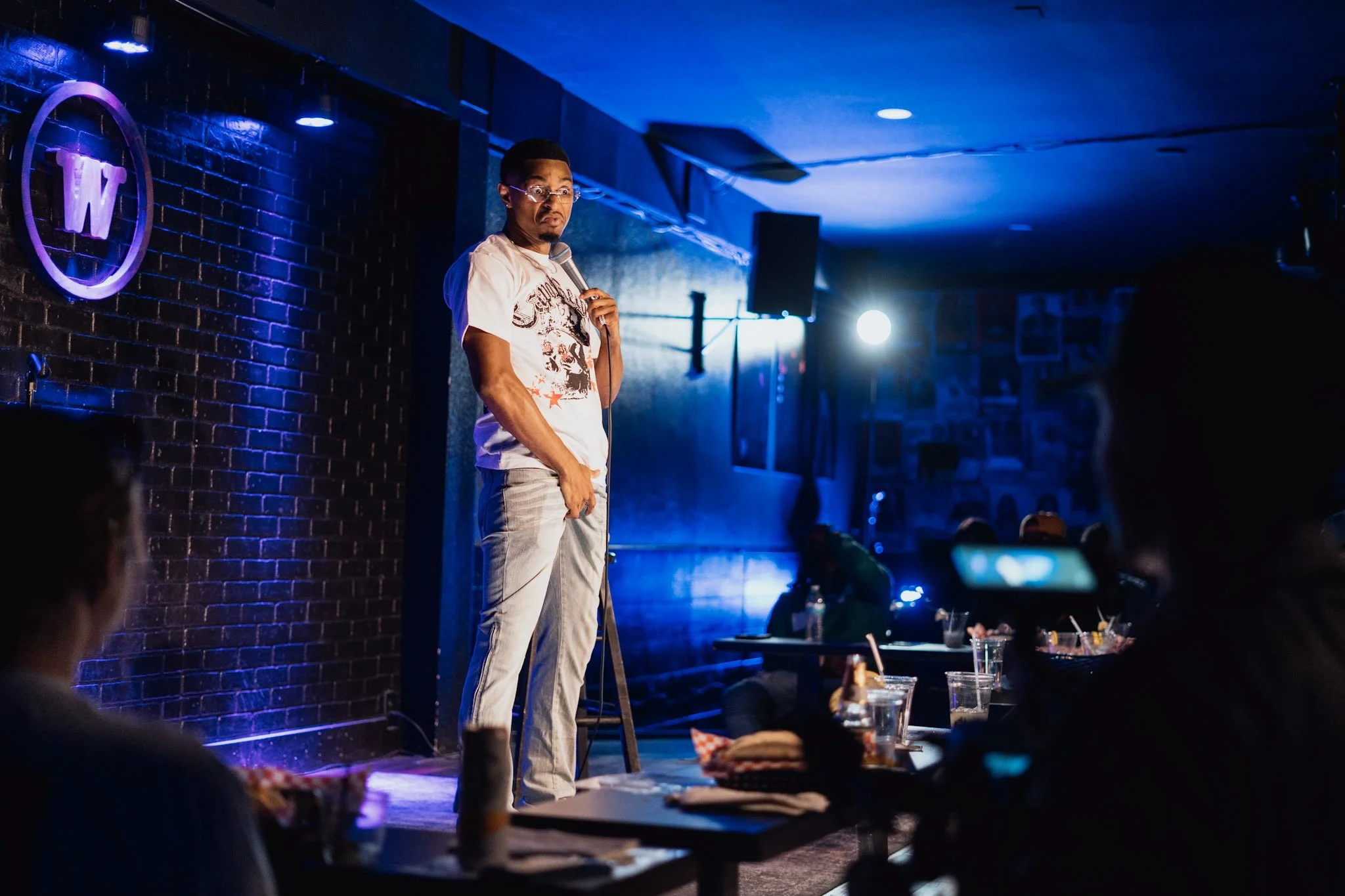 A man standing on stage holding a microphone in a comedy club, with an audience seated at tables in front of him, dim blue lighting, and a brick wall backdrop.