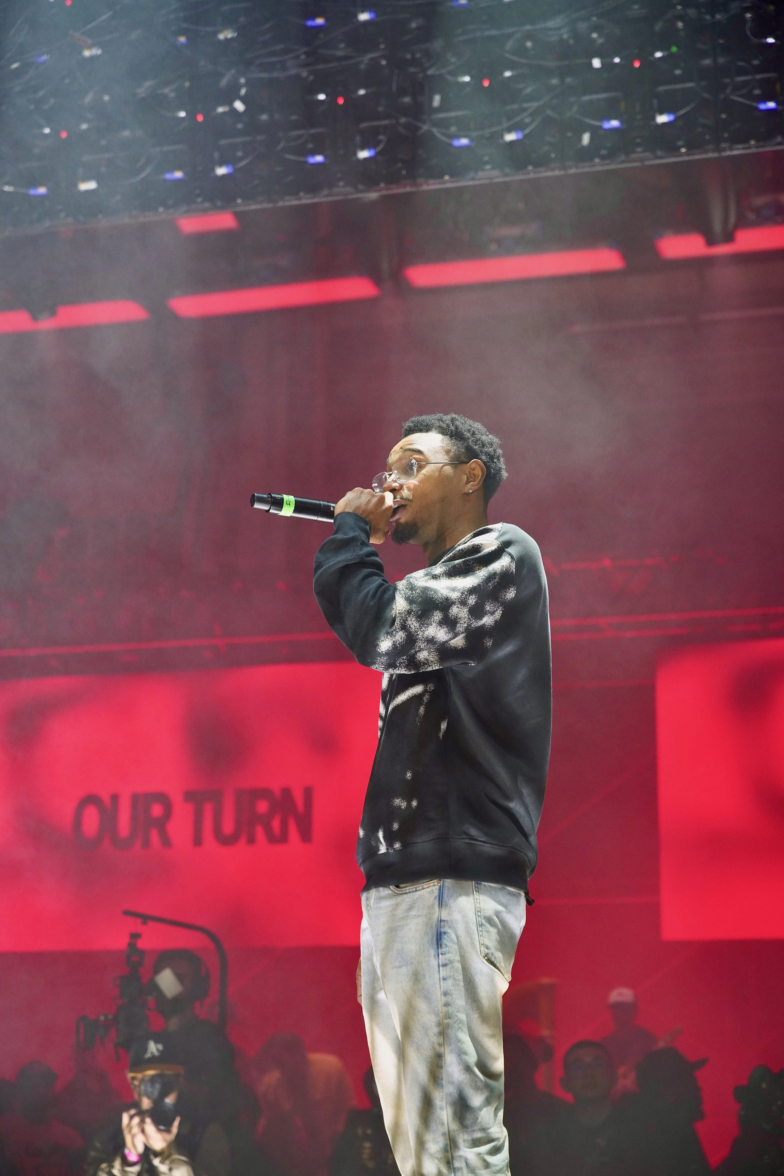 A young man is performing on stage holding a microphone, wearing glasses, a black sweatshirt with white paint splatter design, and light-colored jeans. Behind him is a red backdrop with the words 'OUR TURN' displayed, and there is an audience and sta