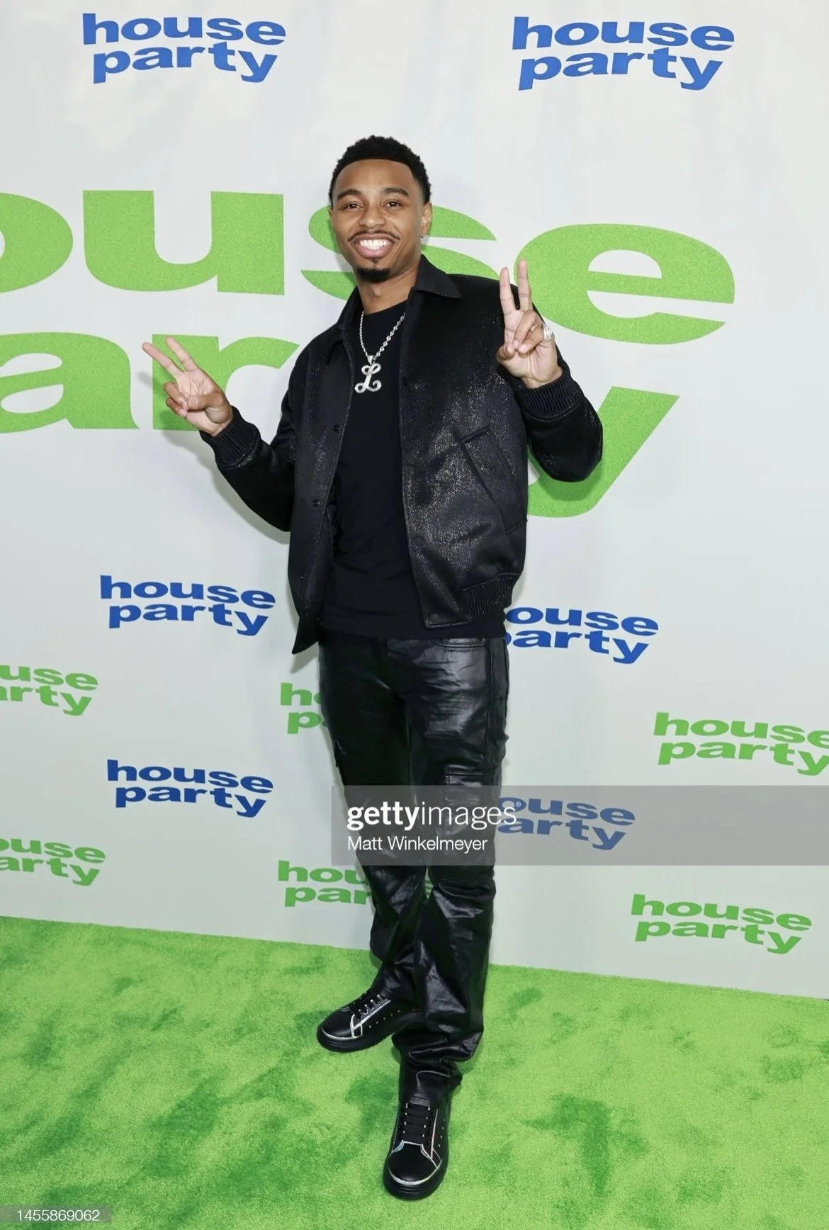 A man smiling and posing with peace signs in front of a backdrop with repeating 'house party' logos, on a bright green carpet.