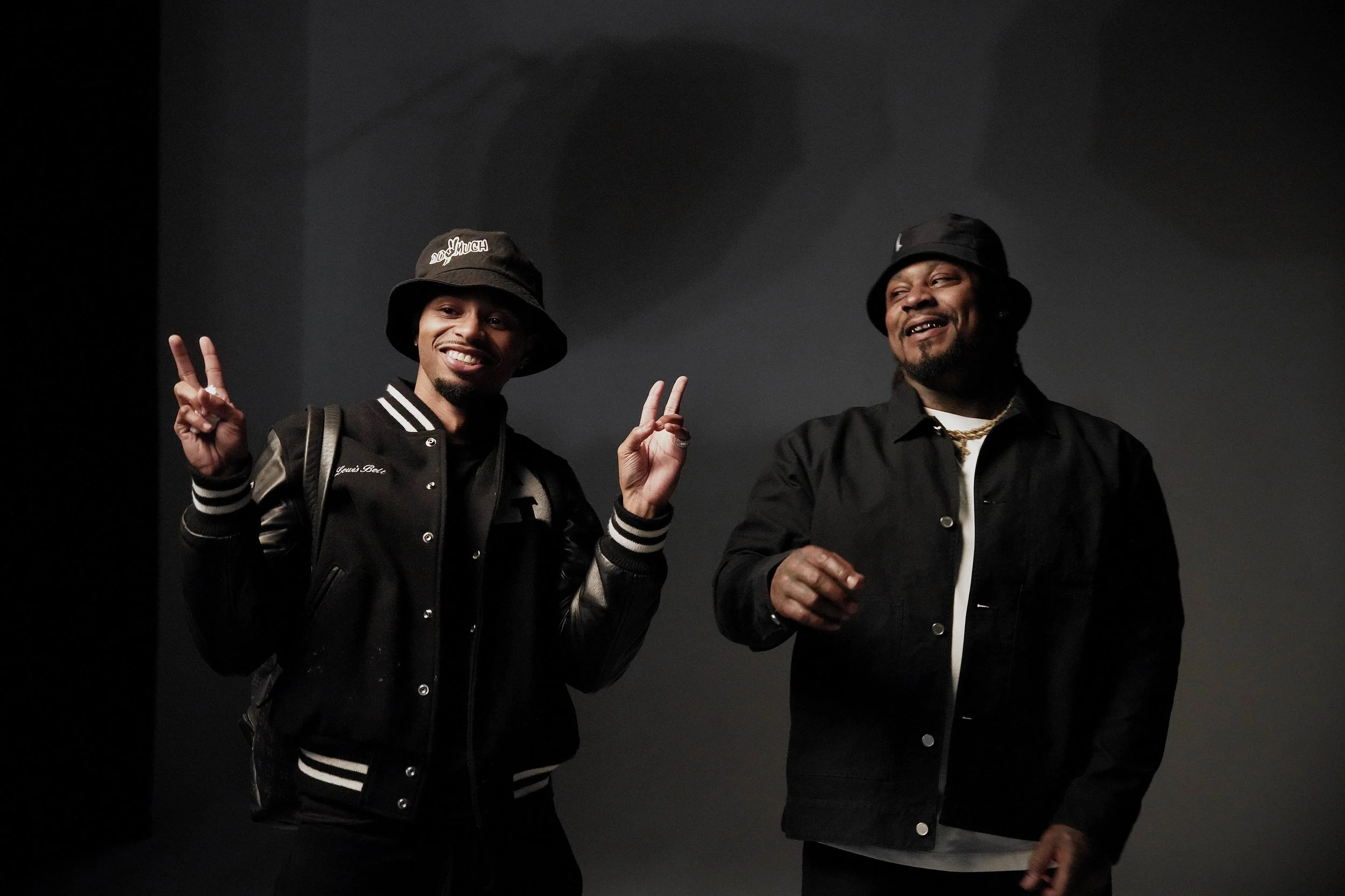 Two smiling men wearing black jackets and bucket hats, making peace signs and standing against a dark background.