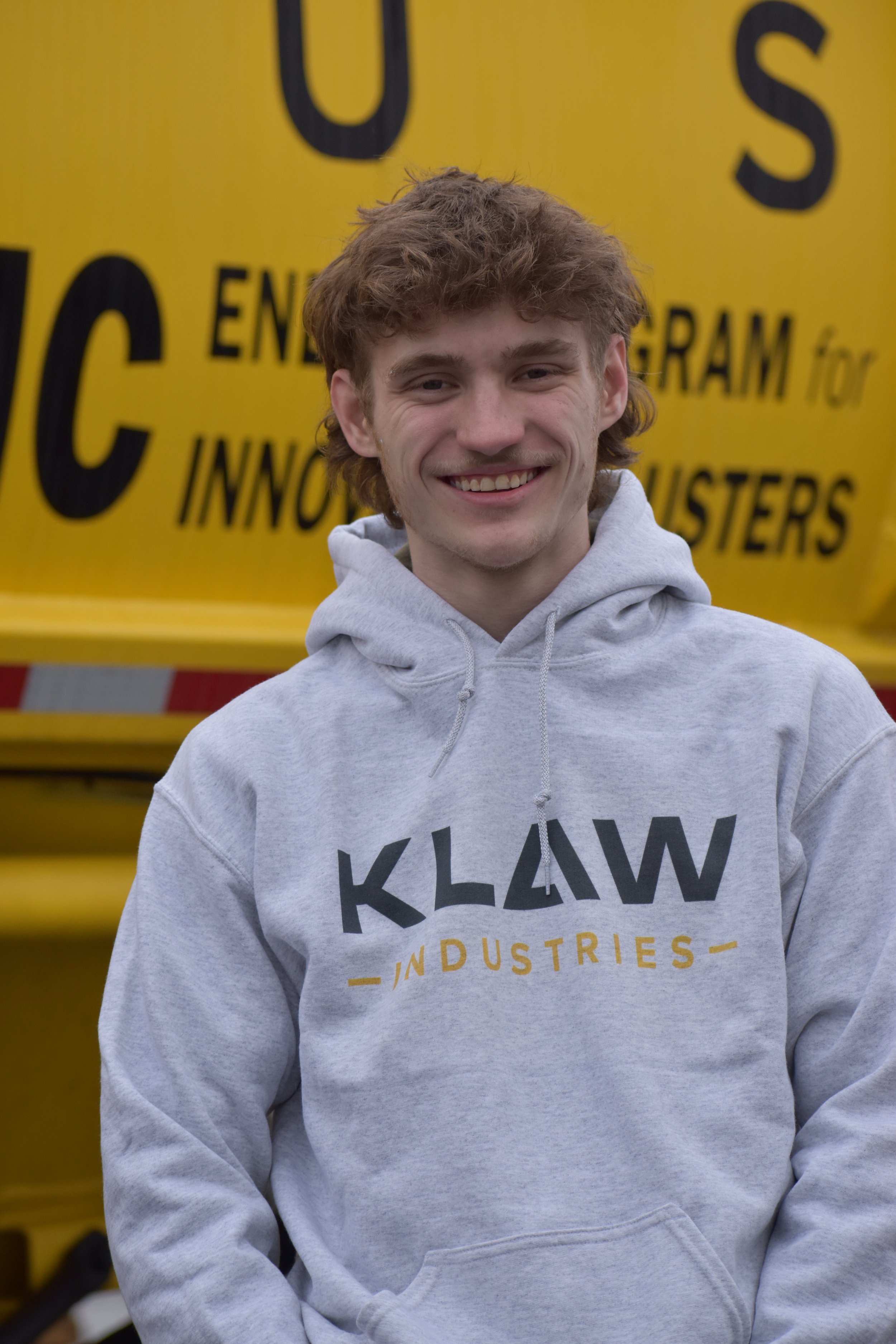 A young man with curly red hair smiling, wearing a gray Klaw Industries hoodie, standing in front of a large yellow vehicle with black text.