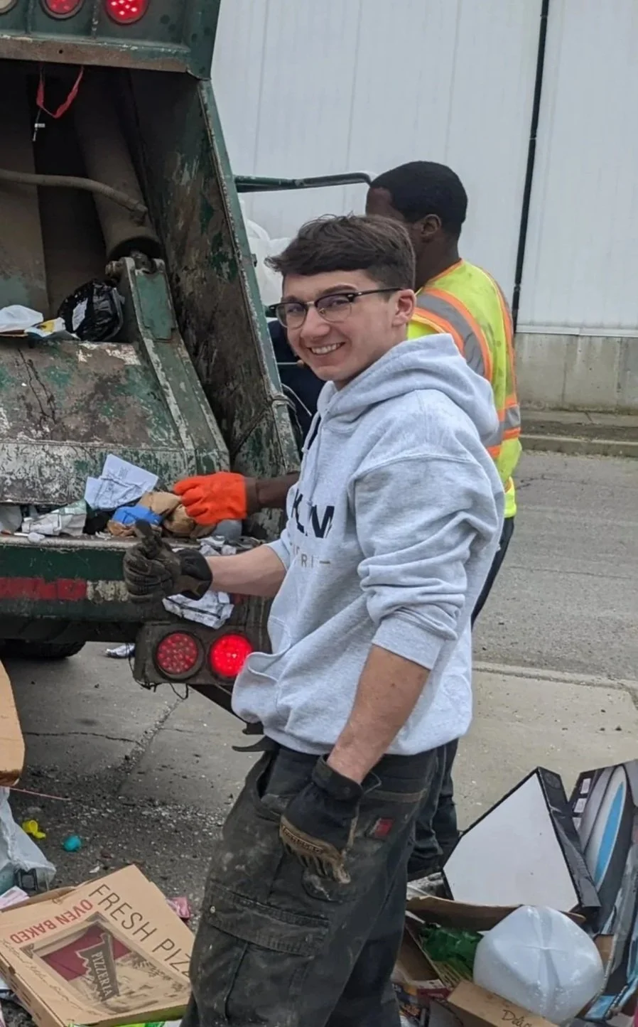 A young man with glasses and a gray hoodie giving a thumbs up, picking up trash near a dumpster with two other men, one in a yellow safety vest, in an outdoor urban setting.
