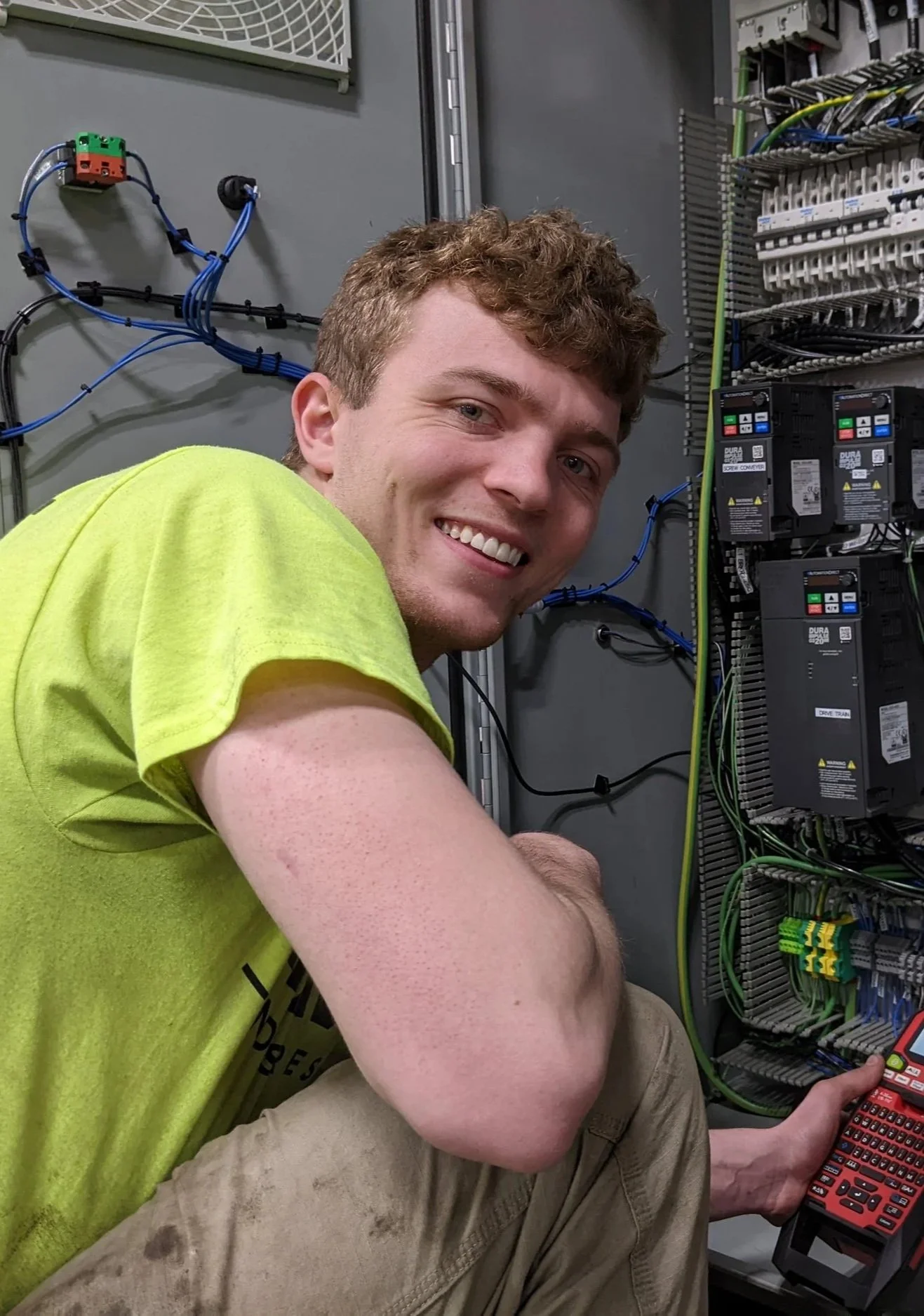 A young man with curly brown hair and a bright smile, wearing a yellow T-shirt, working on an electrical control panel with blue wiring, circuit breakers, and a handheld electronic tester in his right hand.
