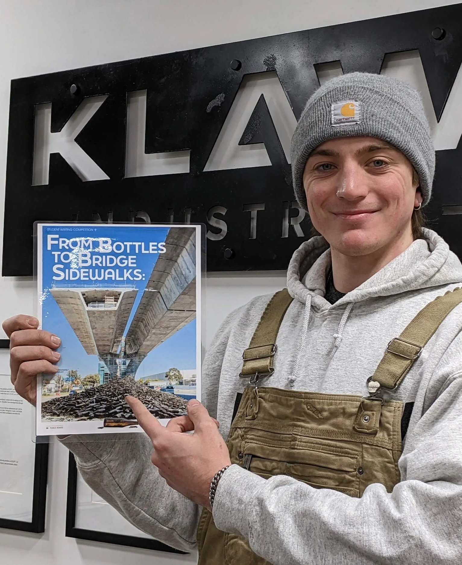 Young man wearing a gray Carhartt beanie and beige overalls pointing at a magazine cover featuring a large bridge and pile of rocks, standing in front of a 'KLAW Industries' sign.