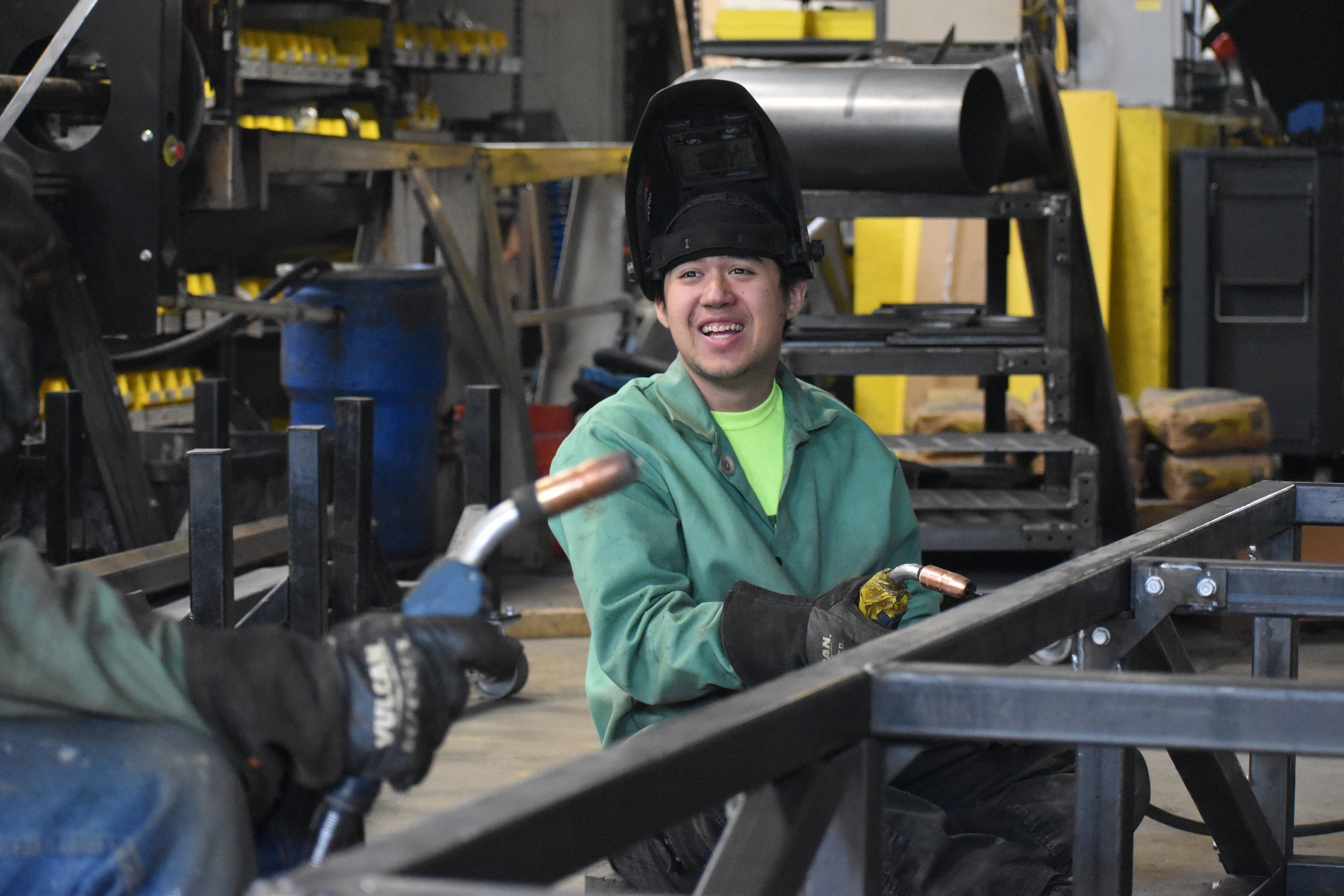 A young man wearing a welding helmet up, green jacket, and gloves, smiling while working on metal frames in a workshop surrounded by tools and industrial equipment.