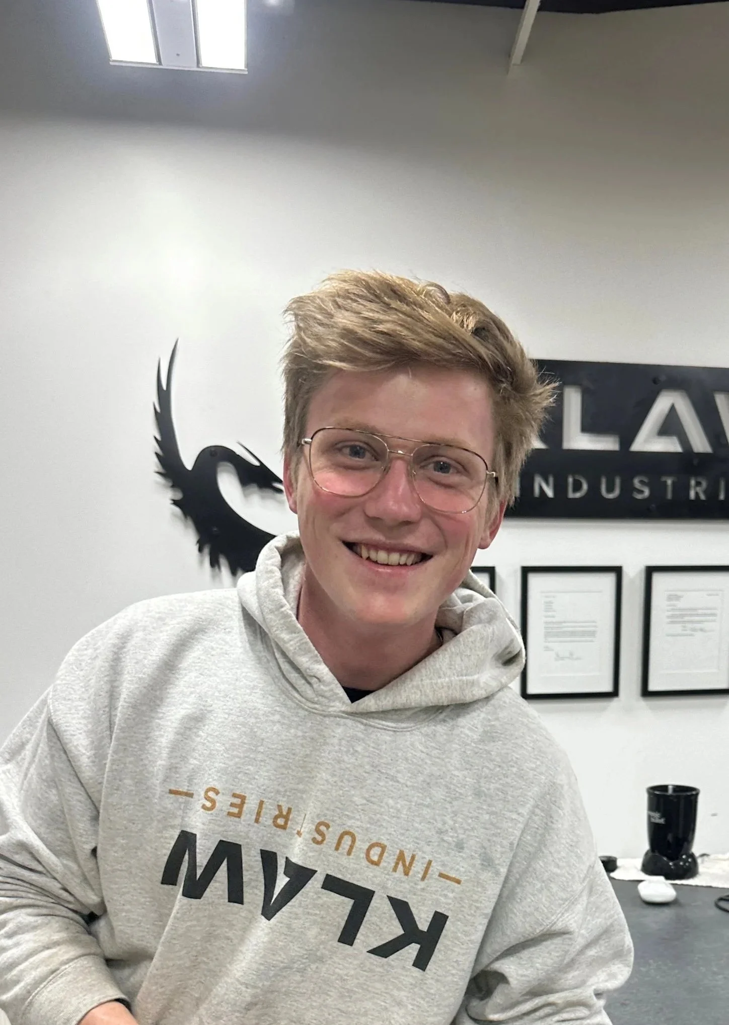 A young man with glasses, blonde hair, and a cheerful smile is wearing a gray hoodie with black and orange text, sitting in an office with a logo and certificates on the wall behind him.