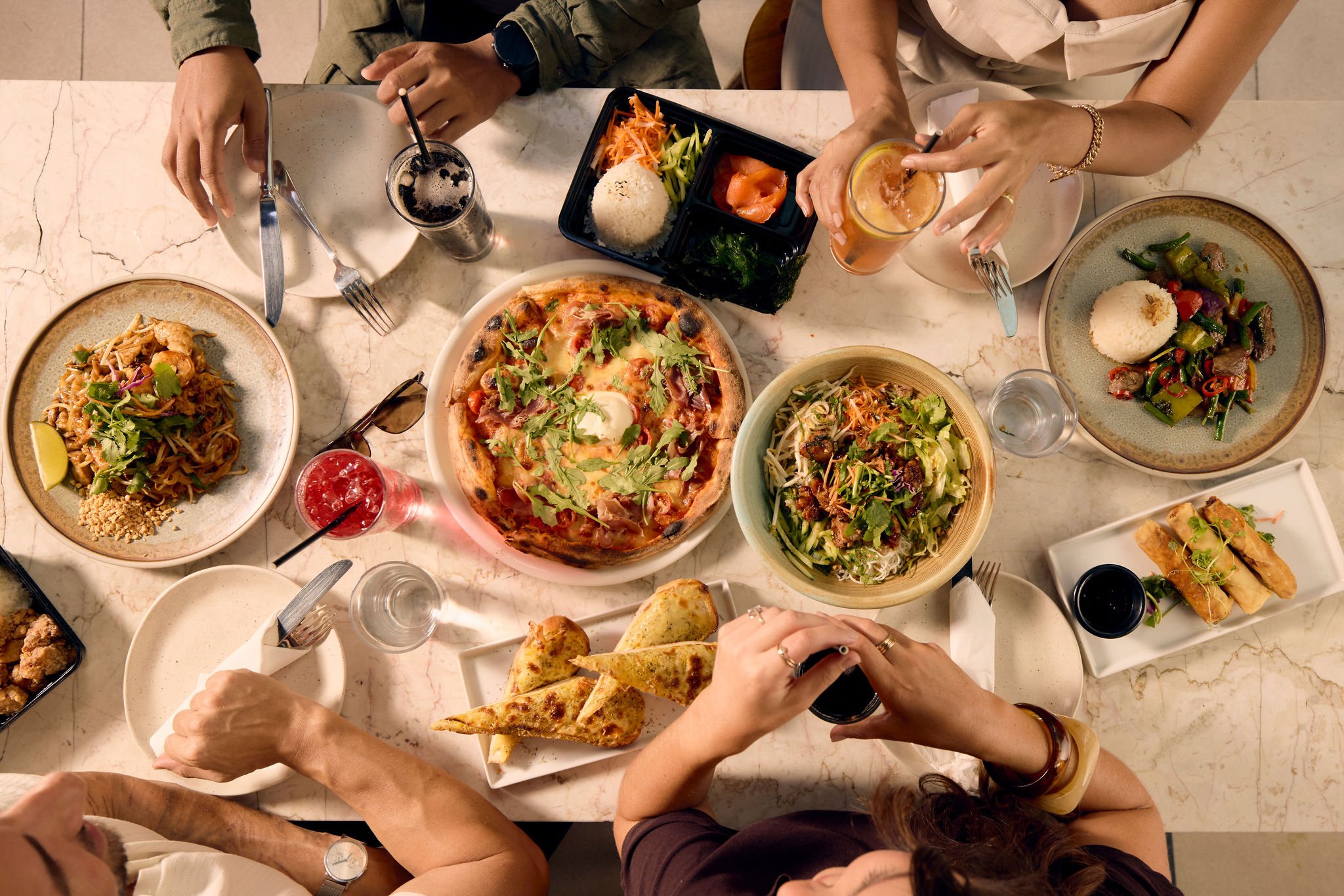 A flatlay spread of various lunch dishes with hands visible. Dishes are pasta, pizza, salad, and some sides like spring rolls