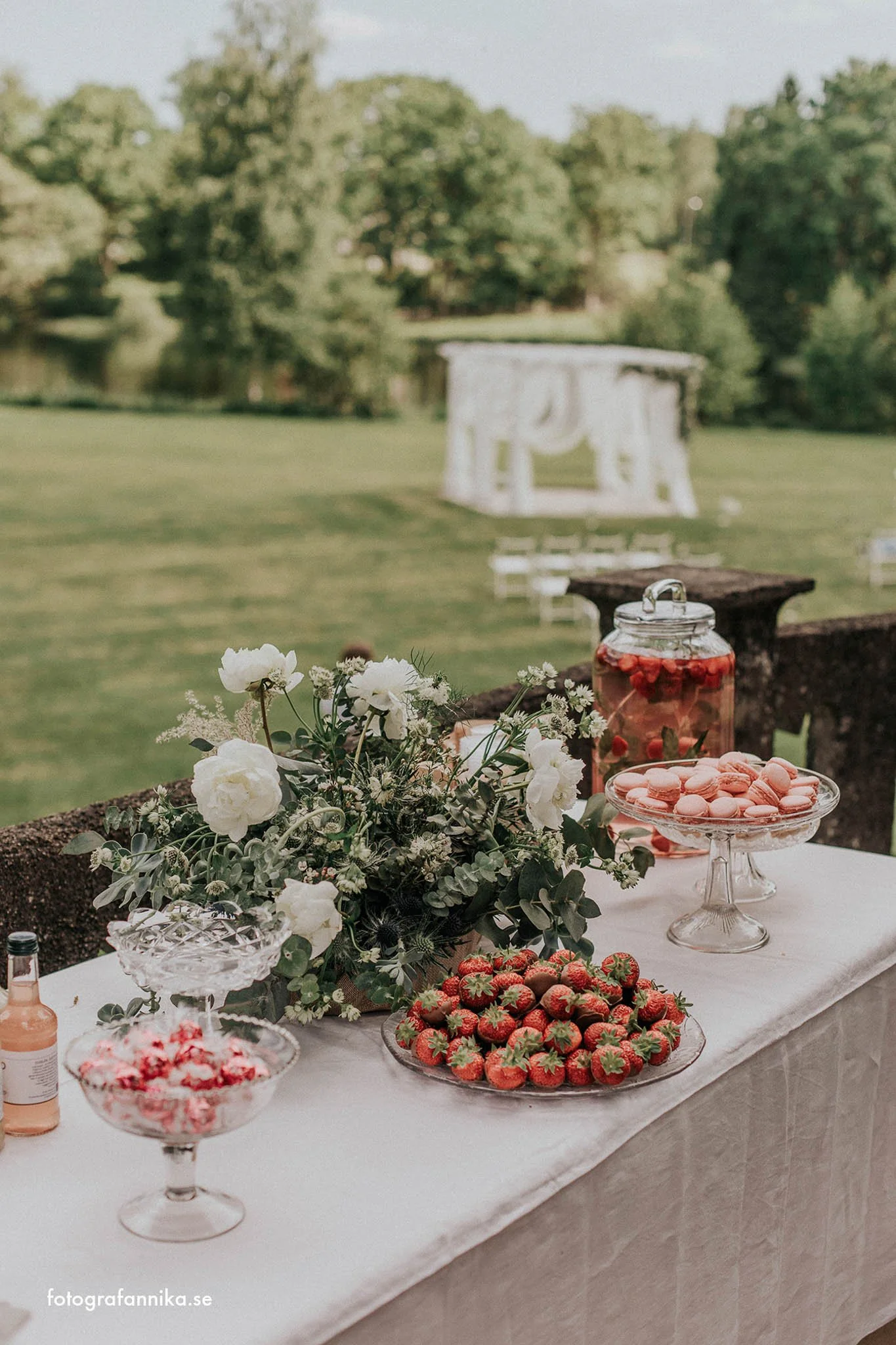 Bord med glasskålar fyllda med jordgubbar och macarons, blomsterarrangemang med vita blommor, utomhuslandskap med grön gräsmatta och träd i bakgrunden, en filt för en fest eller bröllopsfest.