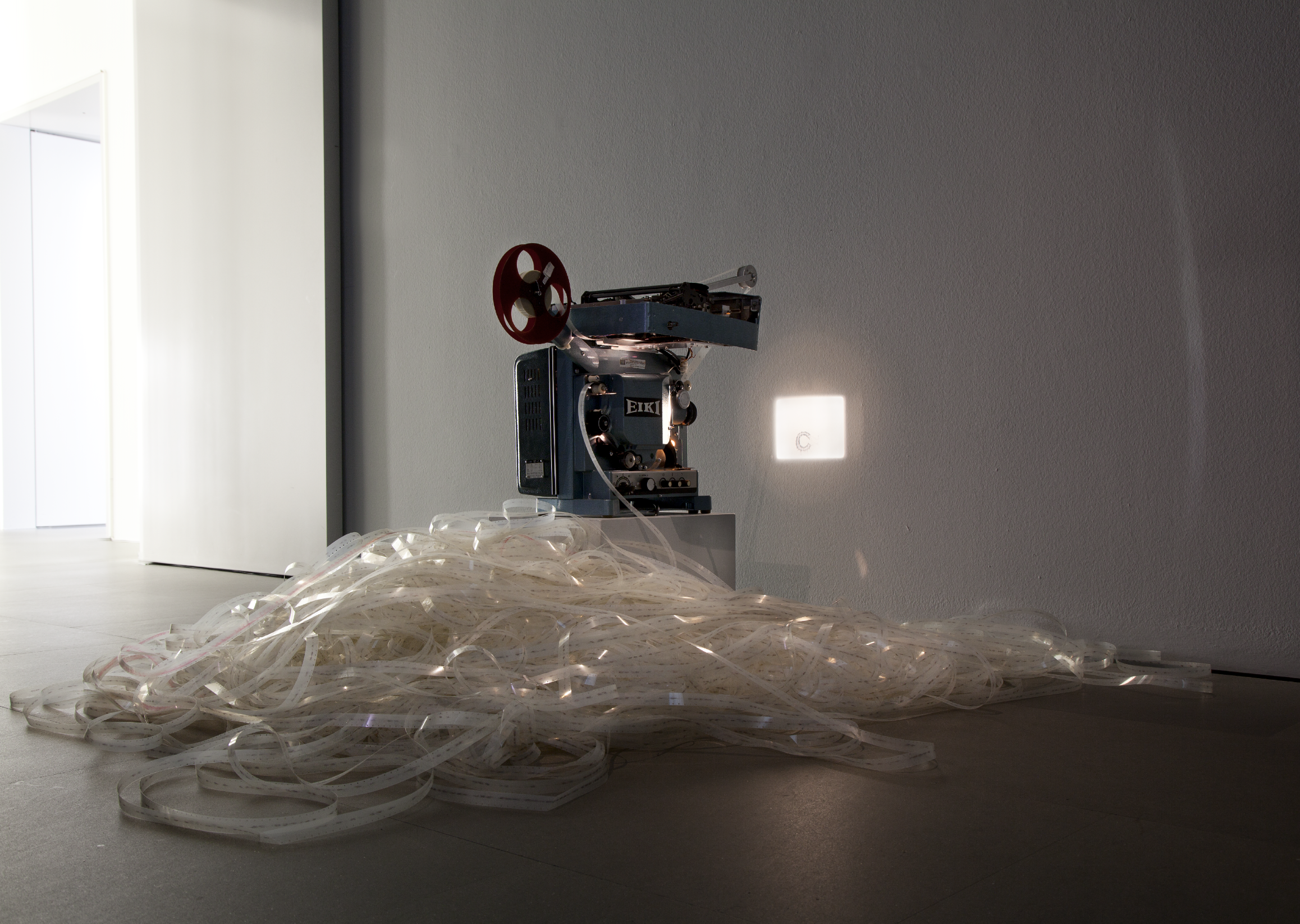 An art installation featuring an old reel-to-reel tape recorder placed on a white pedestal, with a large pile of tangled transparent film strips in front of it, against a plain white wall with a small illuminated square and a door opening in the background.