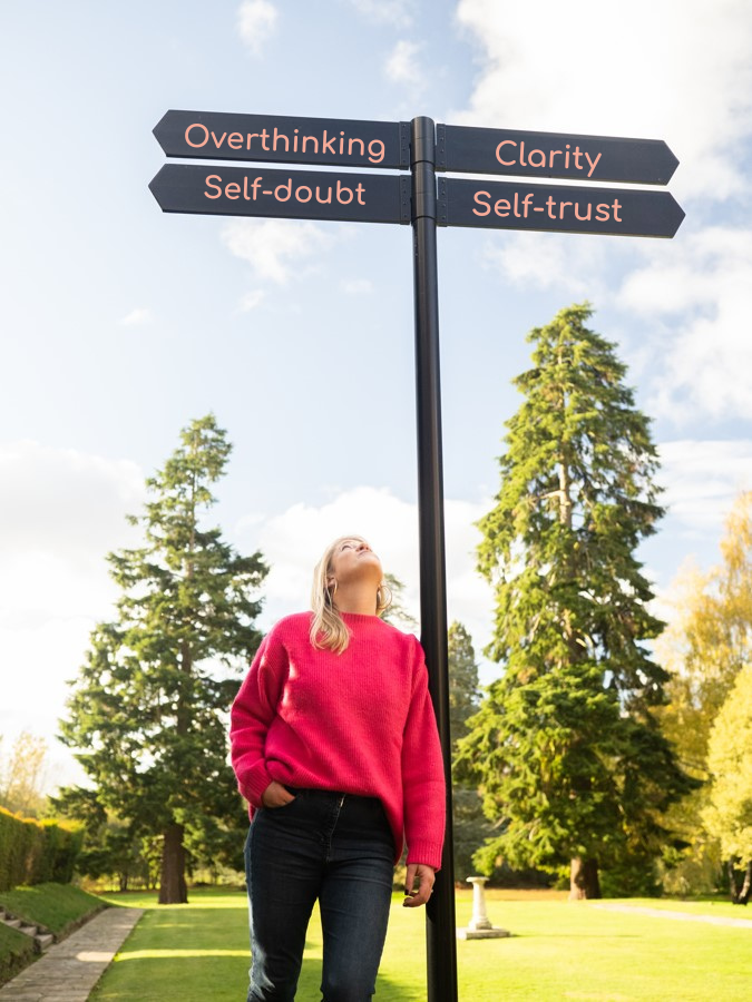 Woman looks up at sign post, overthinking and self-doubt pointing one way, clarity and self-trust pointing the other way.