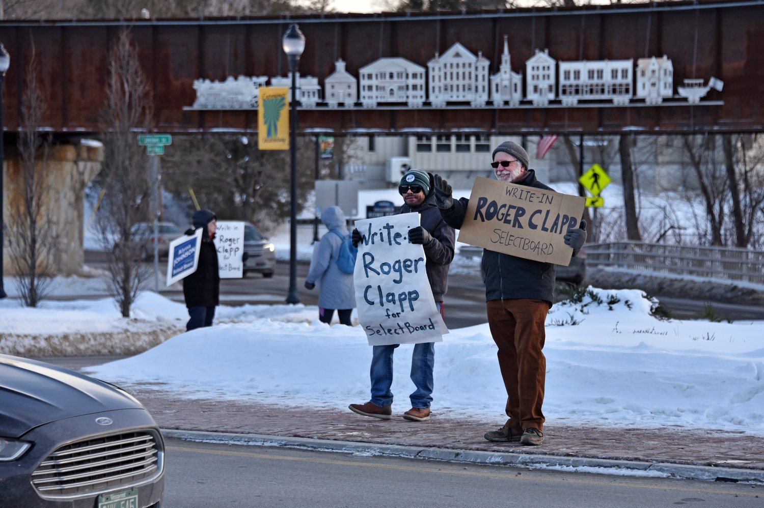   At the roundabout, Maroni Minter, left, and Don Schneider wave hand-painted signs for write-in candidate Roger Clapp on Monday. Photo by Gordon Miller  