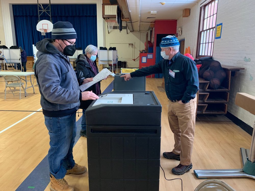   Bob Butler, right, a member of the Board of Civil Authority, directs voters to put ballots in the correct box and machine. Photo by Lisa Scagliotti.   
