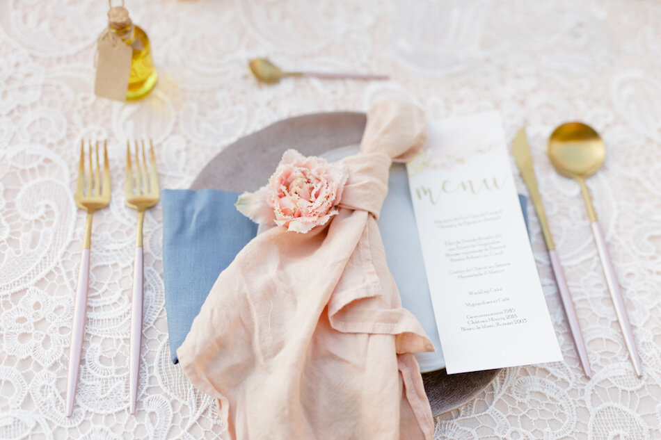 La vaisselle du mariage et le menu, avec une fleur rose claire posée sur l'assiette et des couverts dorés.