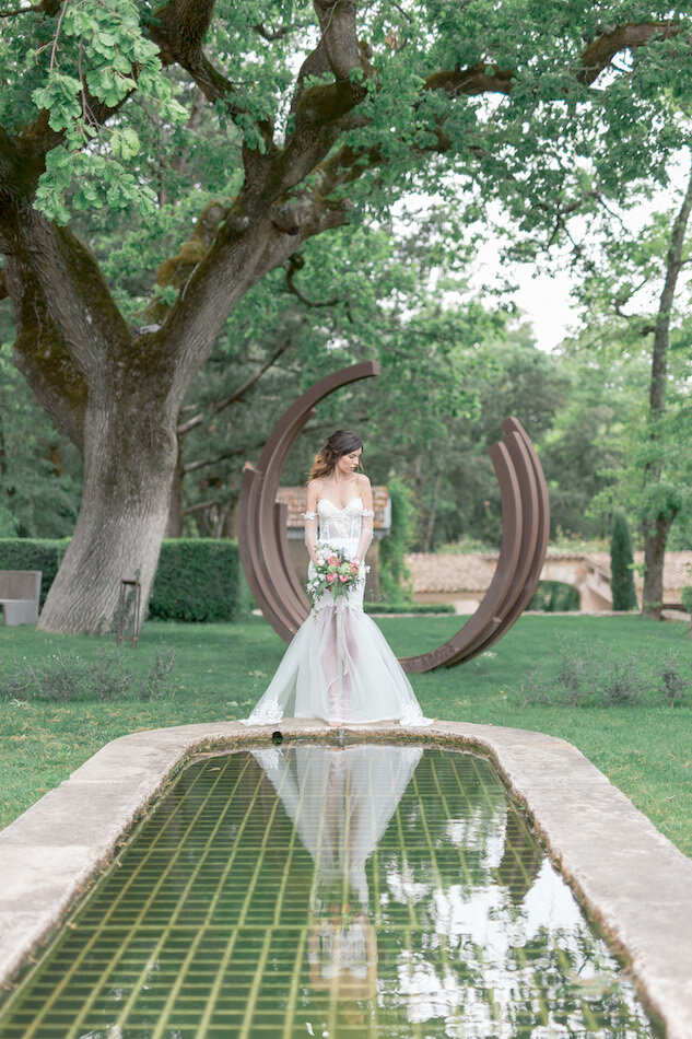 Mariée tenant son bouquet de fleurs devant une fontaine avec son reflet dans l'eau.