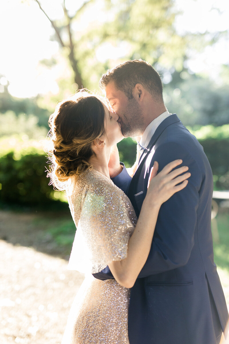 Le couple s'embrassant pendant le first look. La mariée porte une robe fluide blanche pailletée, et le marié un costume bleu marine.