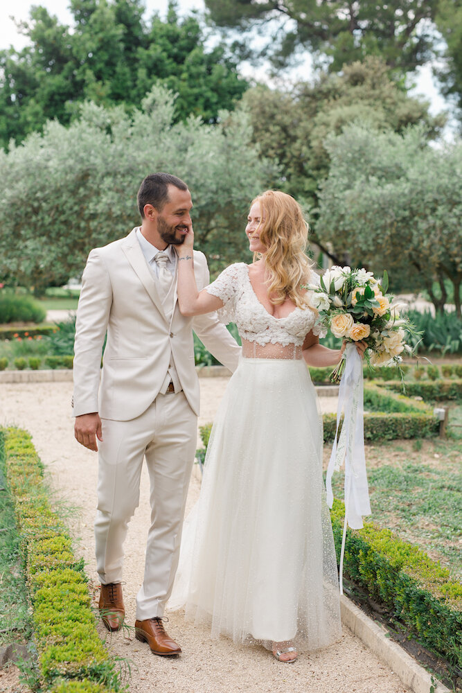 La mariée caressant la joue de son mari lors du shooting couple au mas de la rose en provence, le regardant avec amour et marchant avec lui dans une petite allée du jardin
