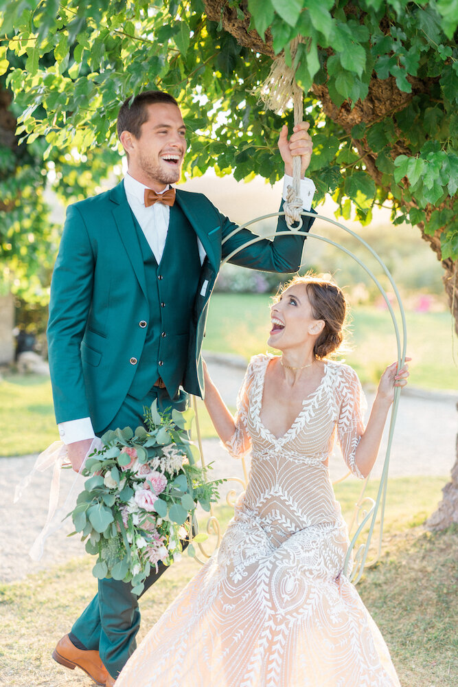 The groom and the bride are laughing out loud together, the bride is sitting on a swing.