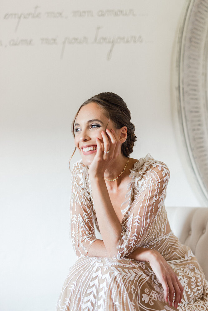 The bride is sitting on a beige armchair and she is smiling and looking outside.
