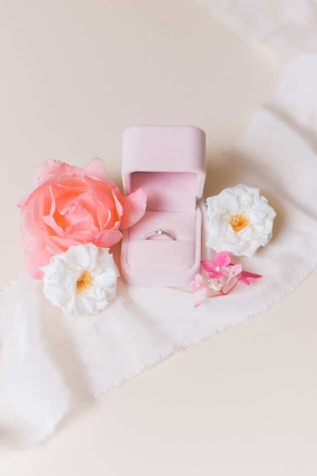 Alliance de la mariée dans son écrin rose poudrée, entourée de fleurs roses et blanches.