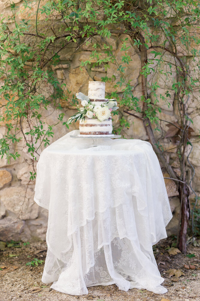 Au centre la table avec nappe blanche et au milieu le naked cake du mariage, blanc et garni de fleurs blanches.