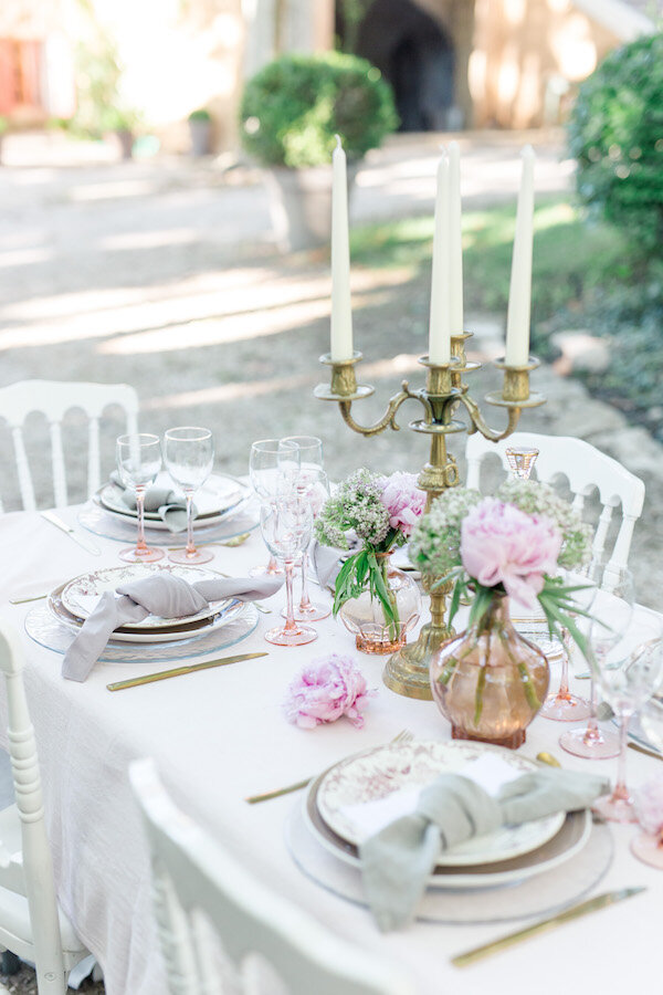 Détails de la décoration de table avec un grand chandelier, des assiettes vintages, des vases de pivoines, et des serviettes grises claires.