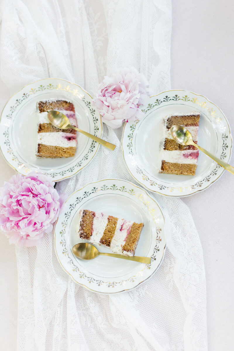 Vue flatlay du dessus de trois assiettes vintage blanches et dorées avec trois parts de wedding cake, entourées de quelques pivoines roses et avec des petites cuillères dorées.