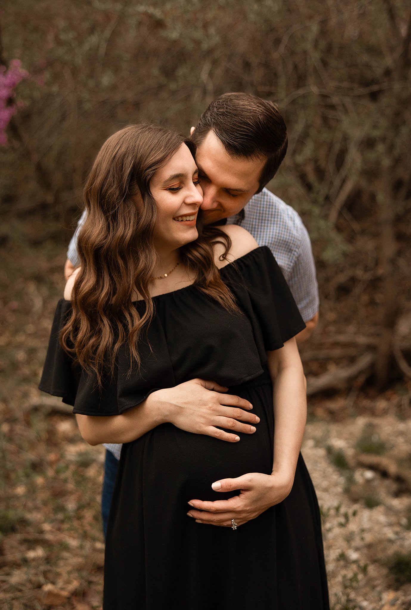 maternity photo of a couple with dad hugging expectant mom from behind while mom holds her belly and smiles