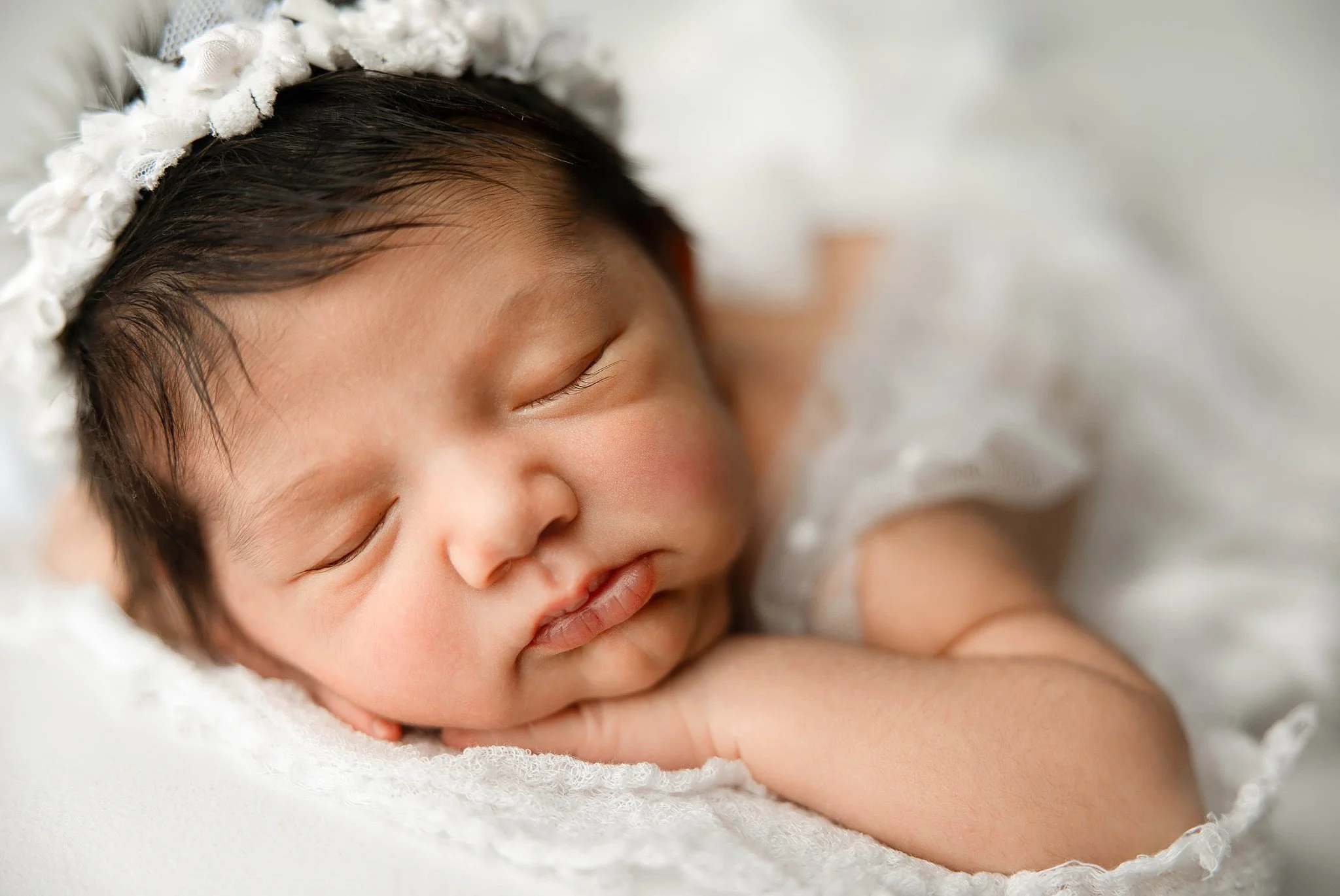 two week old baby girl on a soft white blanket posing with her hands under her chin with a lacey white romper on and matching headband