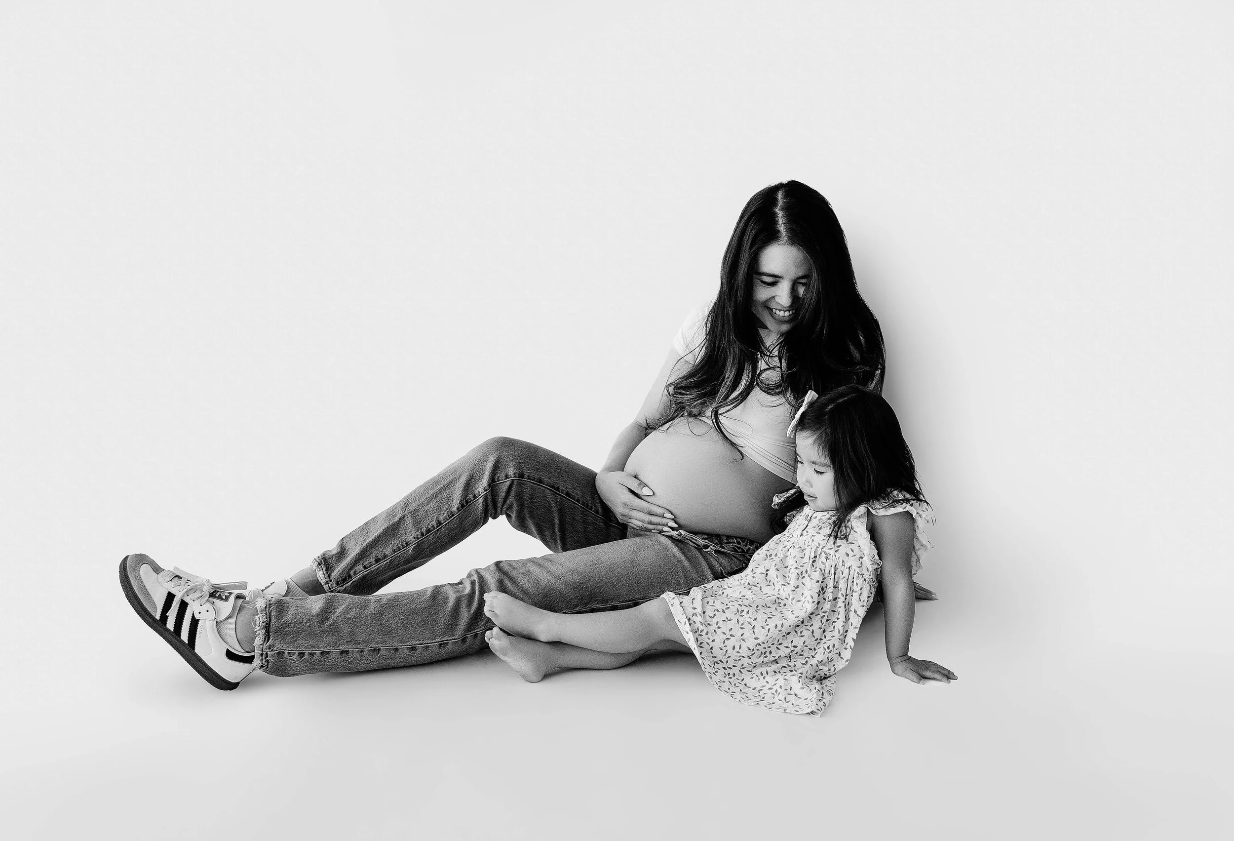 instudio maternity picture of mom with her baby bump showing while she holds onto her belly and smiles sweetly at her toddler daughter in the photo with her