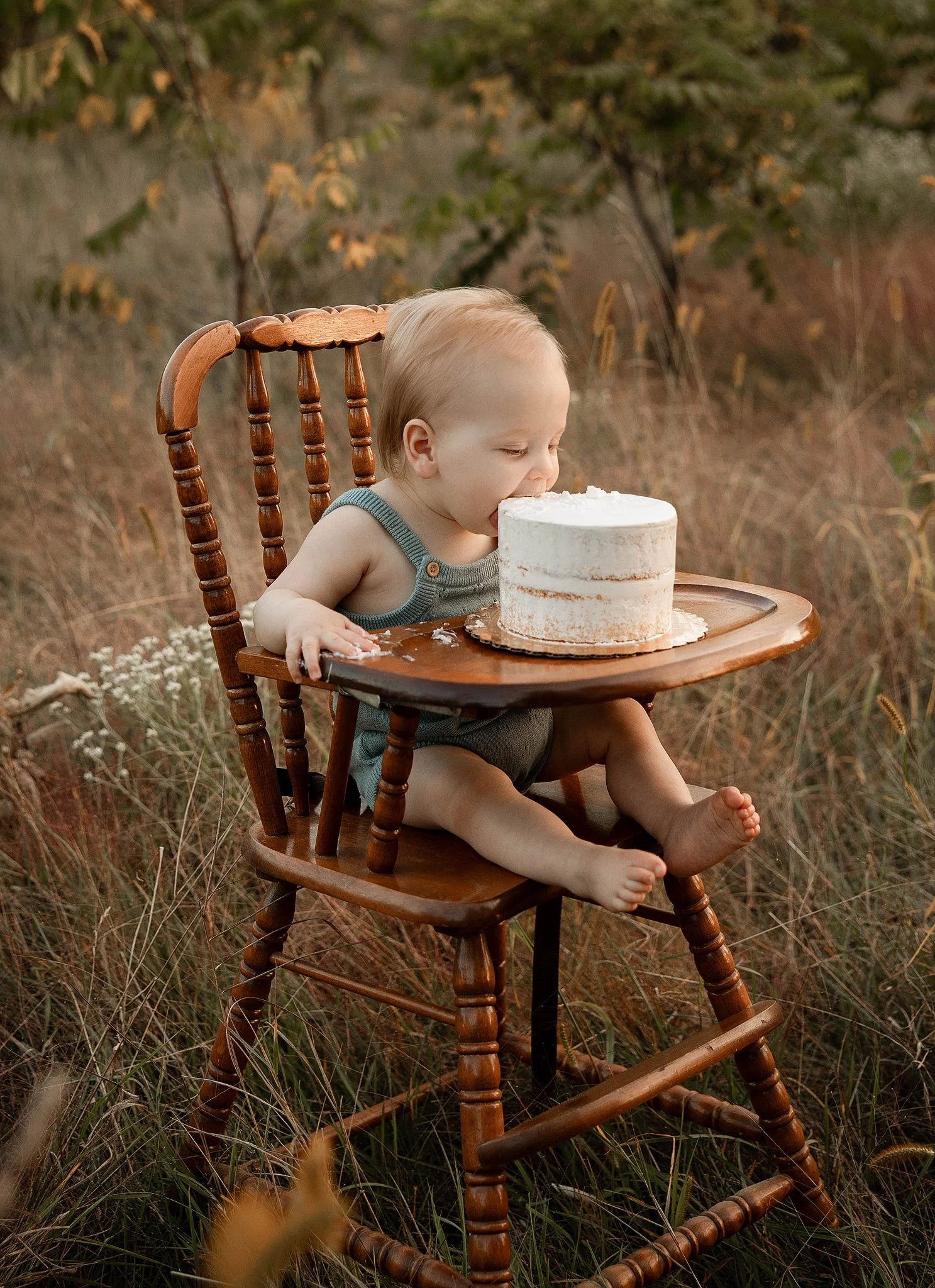 one year old baby boy sitting in a brown vintage high chair wearing  light blue romper for his first birthday with a small smash cake in front of him to eat