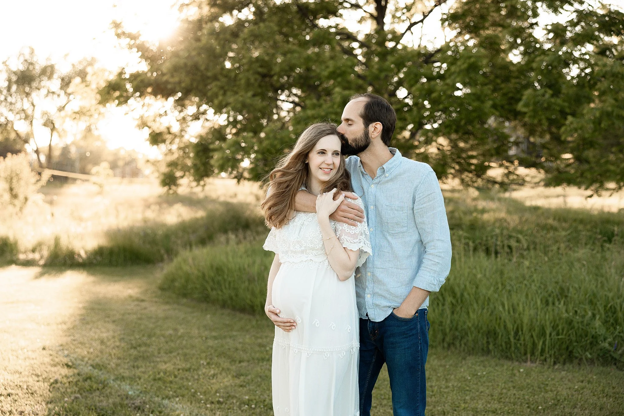 pregnant mom wearing a white lacey dress for her maternity photos with her husband standing behind her and kissing her lovingly on the head at golden hours at a beautiful park with tall grass