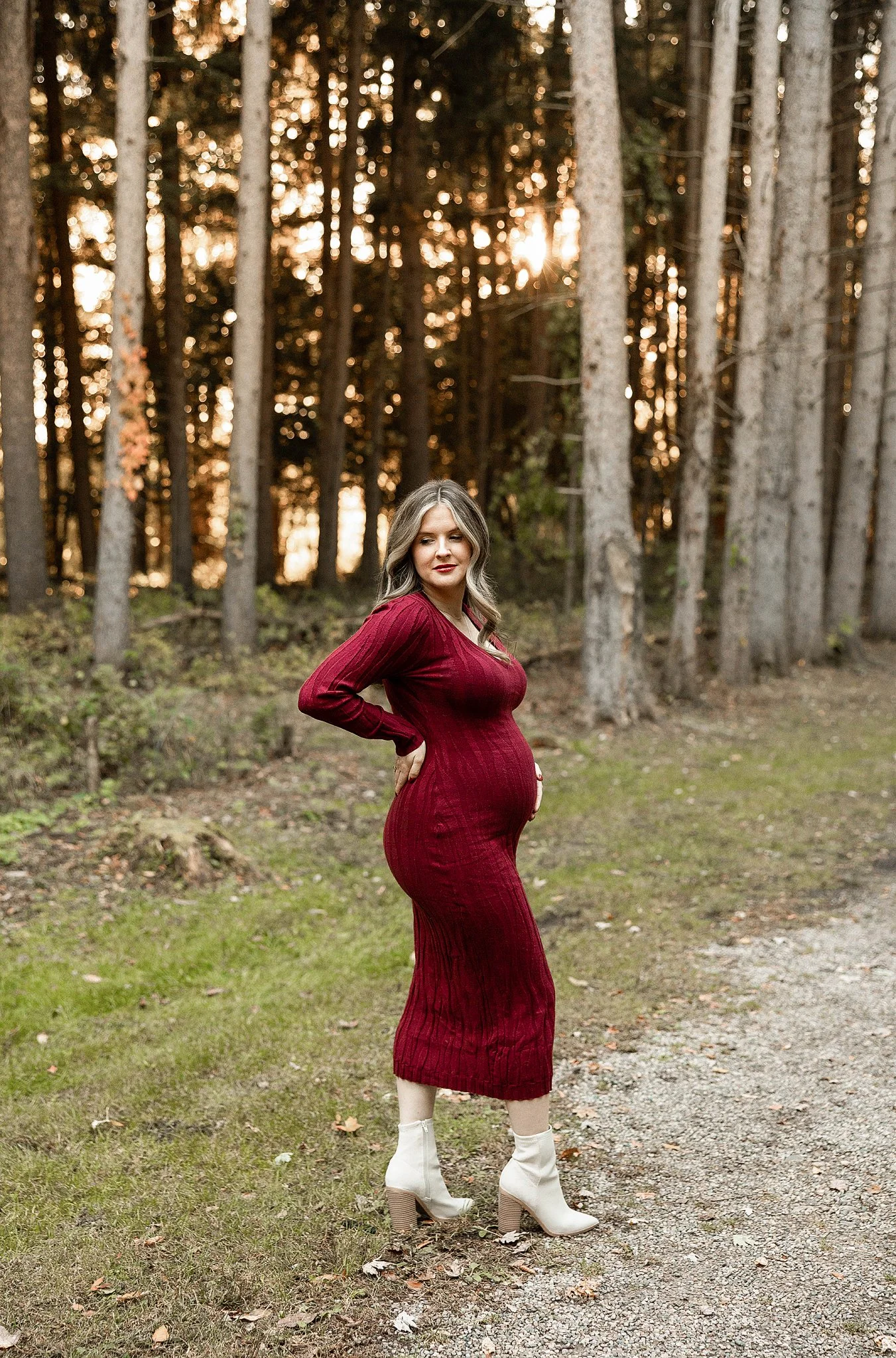 Pregnant mother posing for her maternity pictures wearing a long, form fitting dress and white dressy boots outside at golden hour