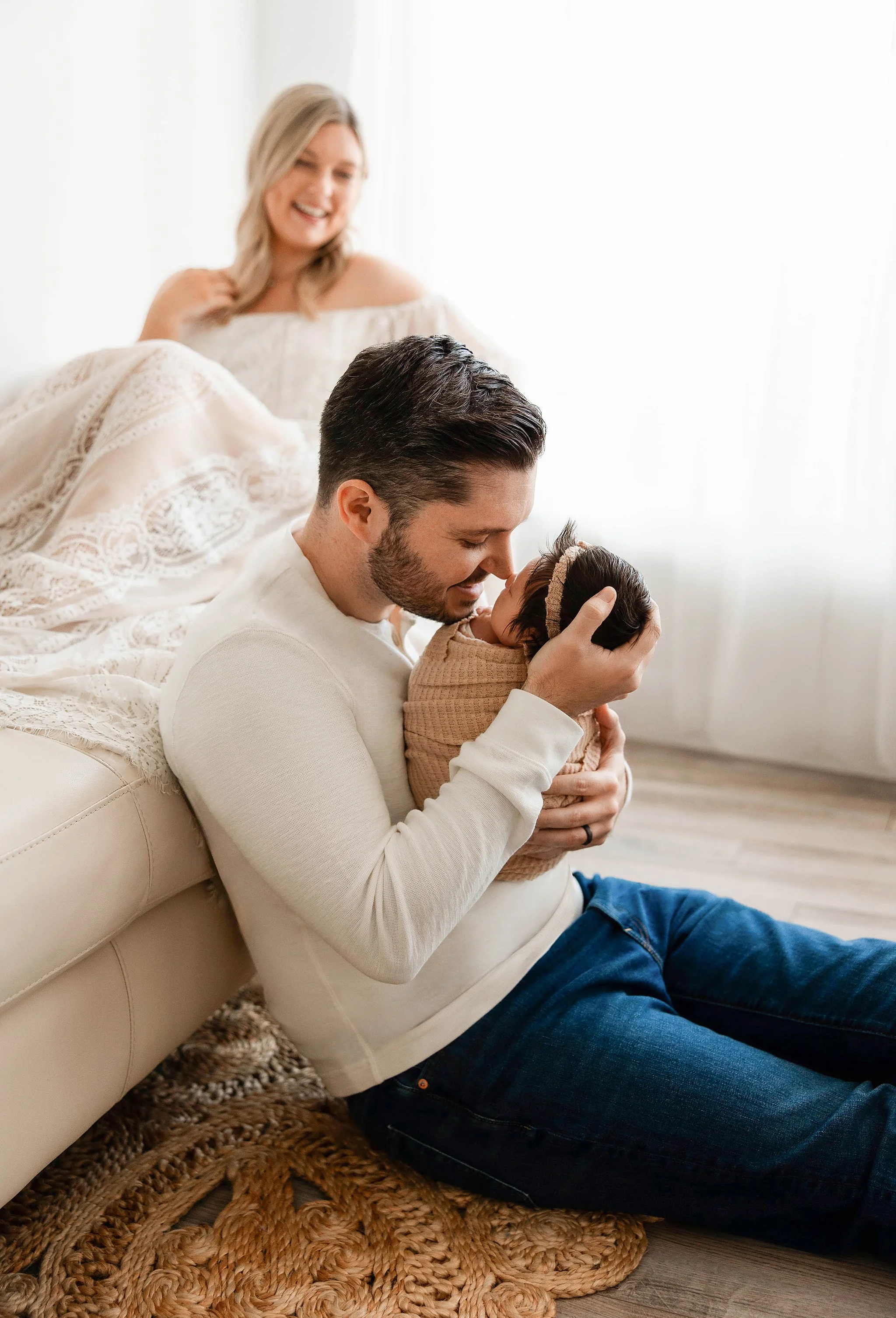 lifestyle family photo of mom on white couch and dad sitting on the floor in front of her holding his baby girl nose to nose in a natural light photography studio
