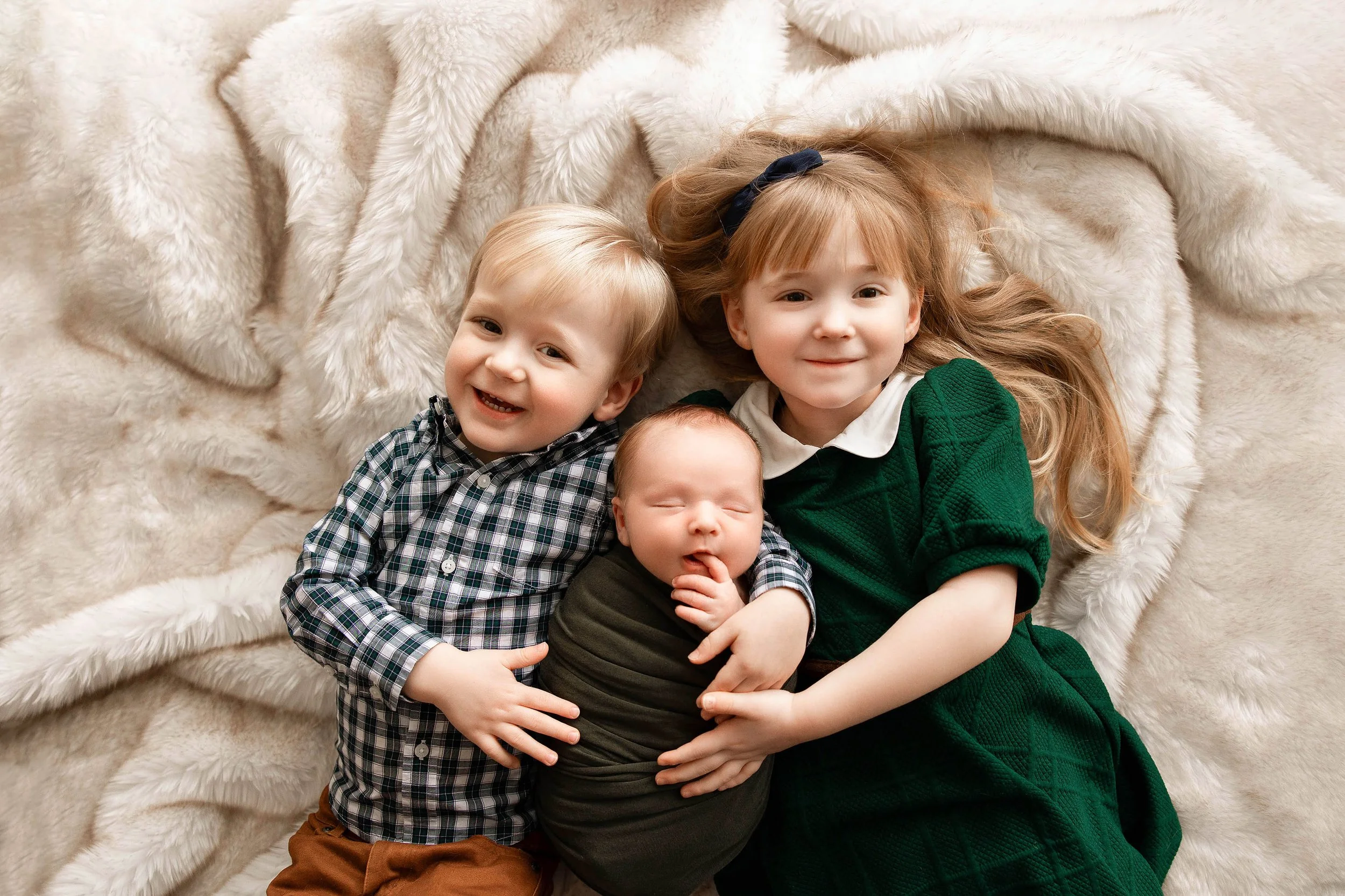 brother and sister holding new baby while laying on soft and comfortable cream color blanket