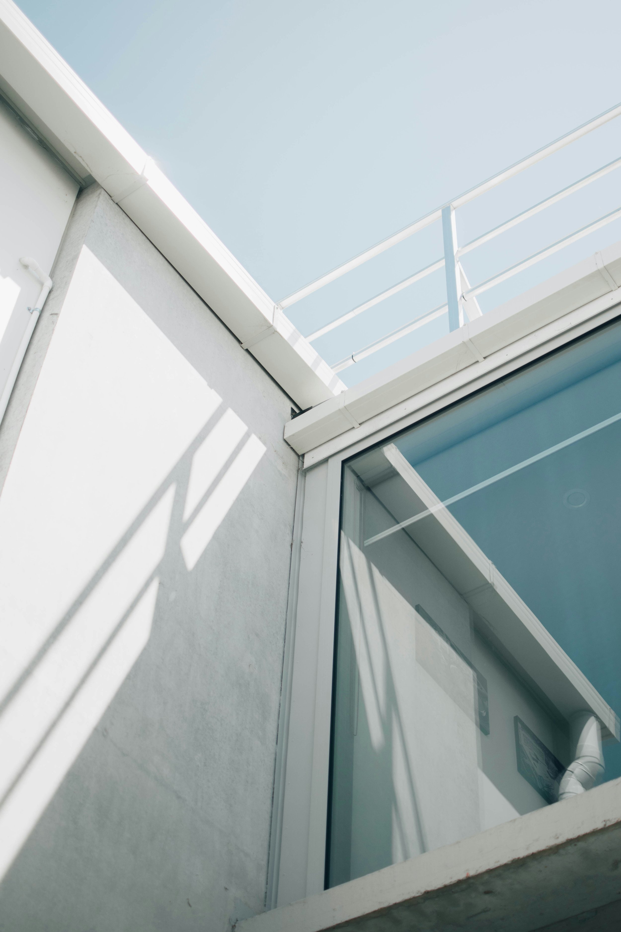 Part of a building exterior showing a white wall, a window with sliding glass doors, and a small balcony with a metal railing. Shadows of the balcony railing are cast on the wall.