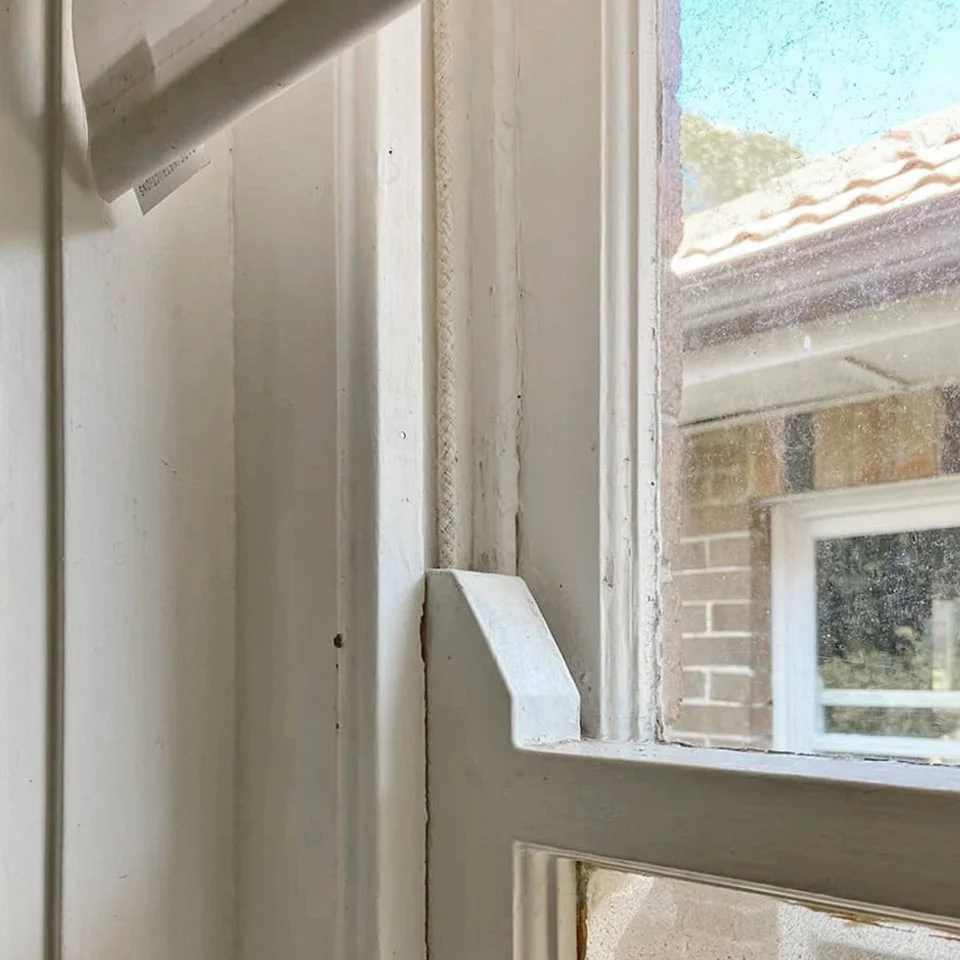 Close-up of a window frame with worn and peeling white paint, with part of the window glass and a brick house visible outside.