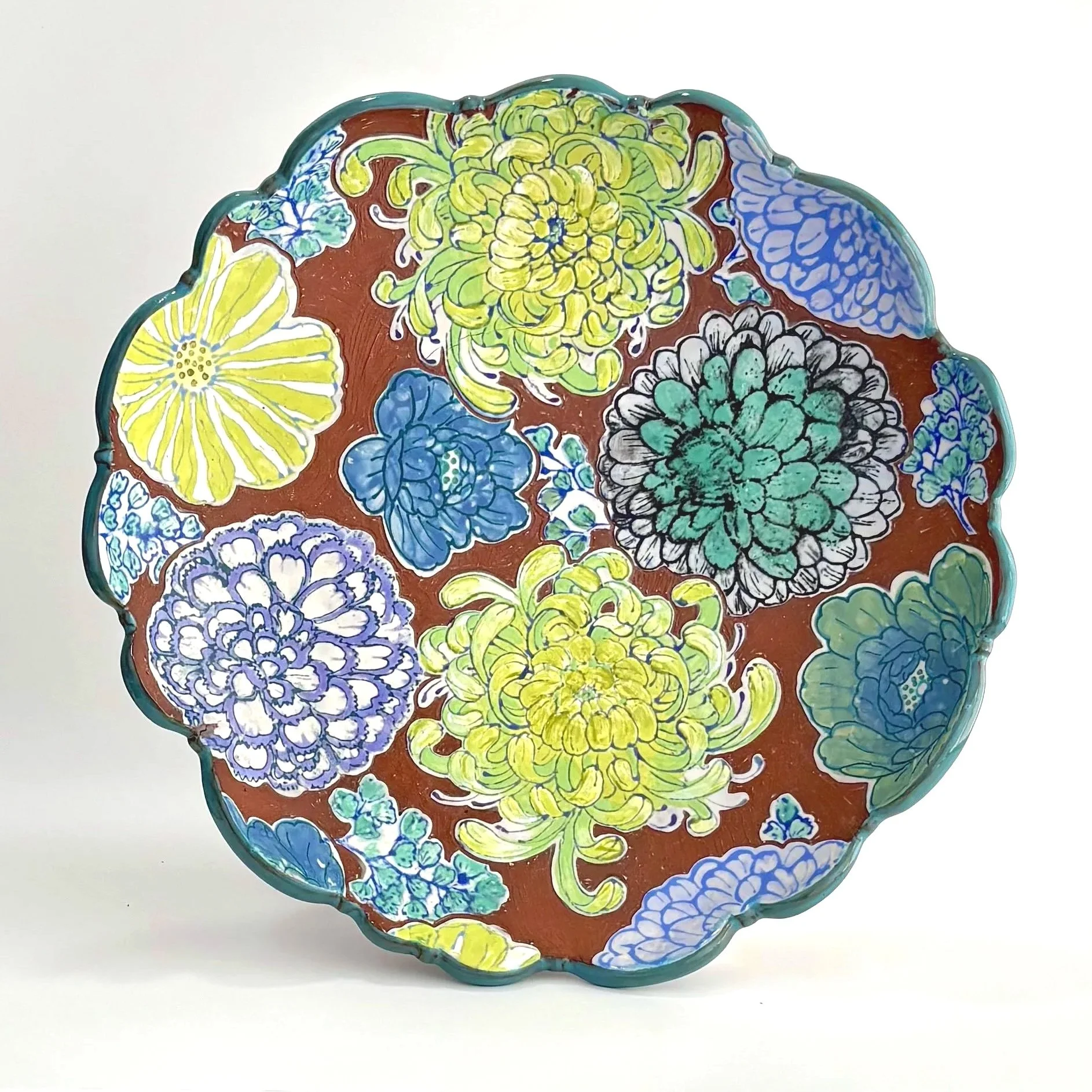 Decorative ceramic plate with a scalloped edge, featuring colorful hand-painted floral patterns in yellow, purple, blue, green, and black on a rust-colored background.