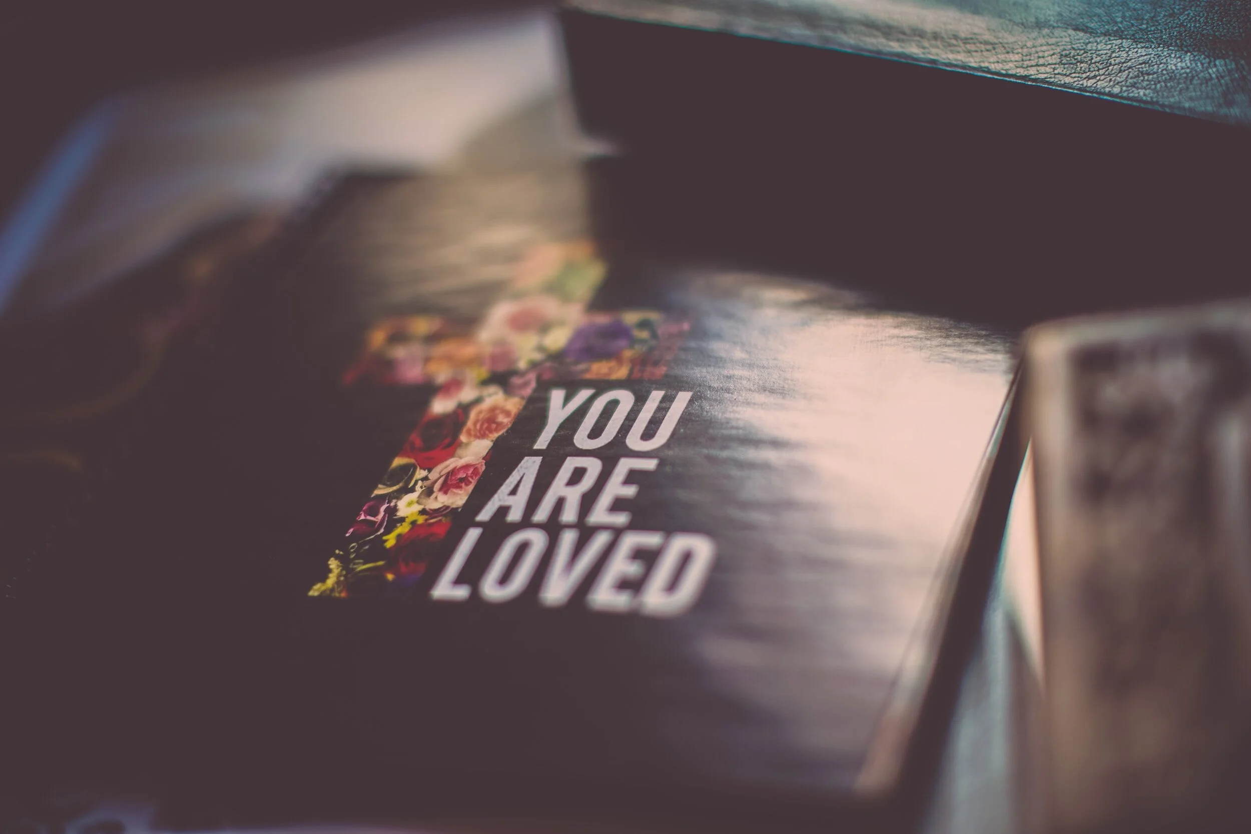 Black book with the words "You Are Loved" on it and a cross with multicolored roses within it on the cover sit on a table or a shelf.