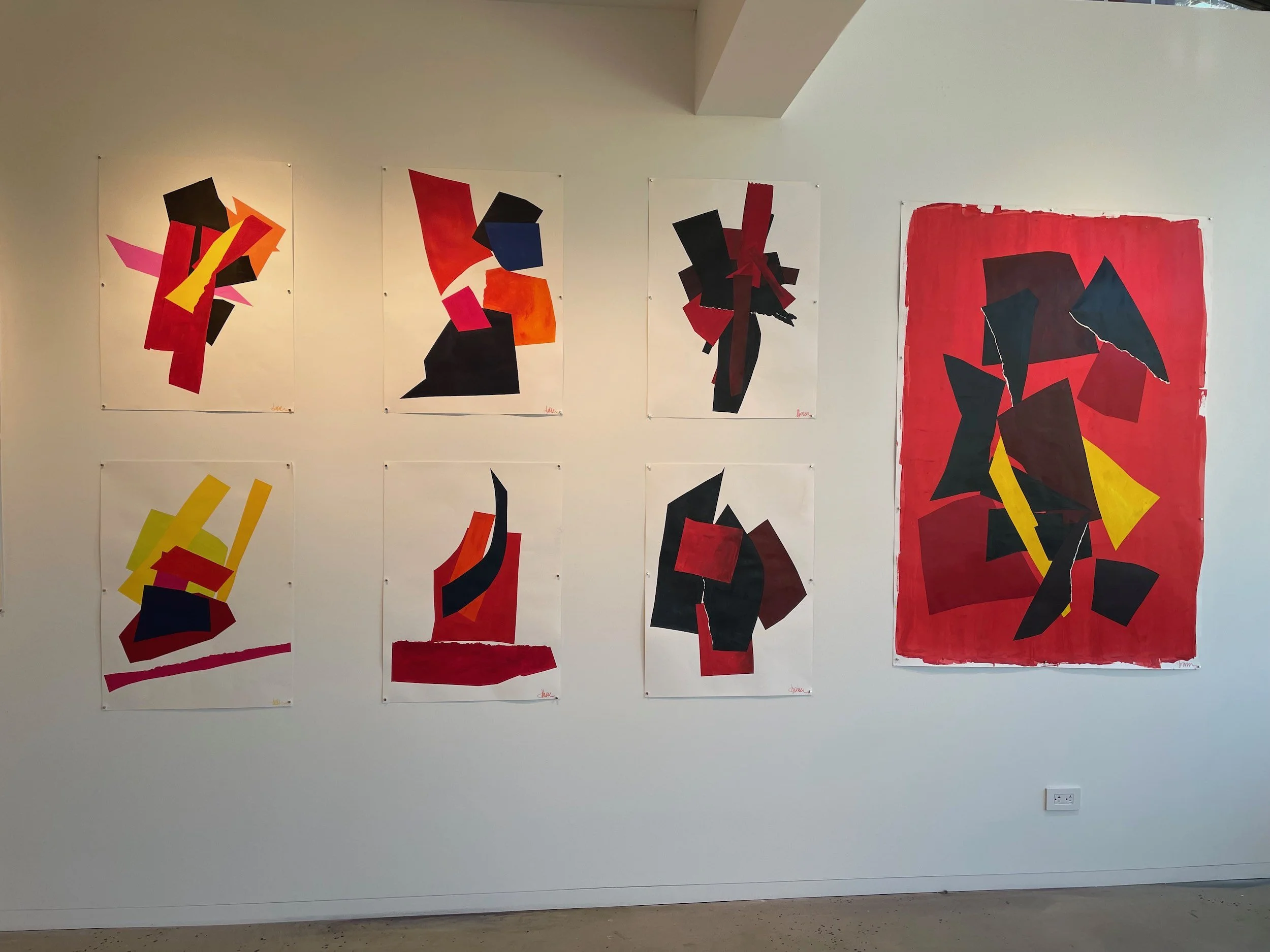 Gallery wall with eight abstract paintings featuring bold geometric shapes in black, red, yellow, and orange, arranged in a grid with seven smaller pieces on white backgrounds and one larger red background piece.