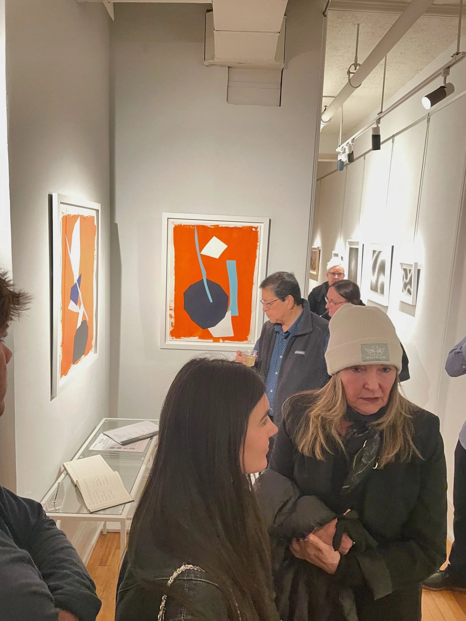 People viewing abstract artwork in an art gallery, with orange, black, white, and blue shapes on the paintings.