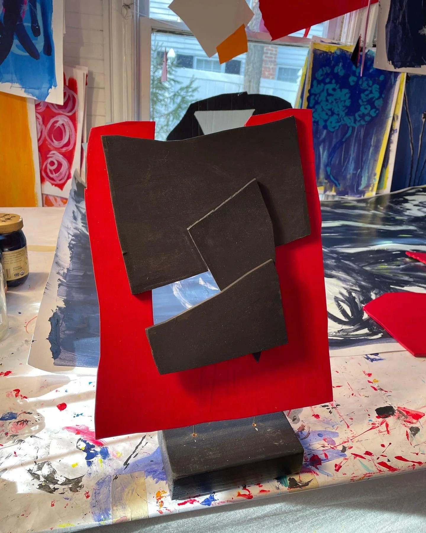 An abstract art sculpture with black rectangular shapes on a red base, painted and displayed in an art studio with colorful paintings and supplies around.