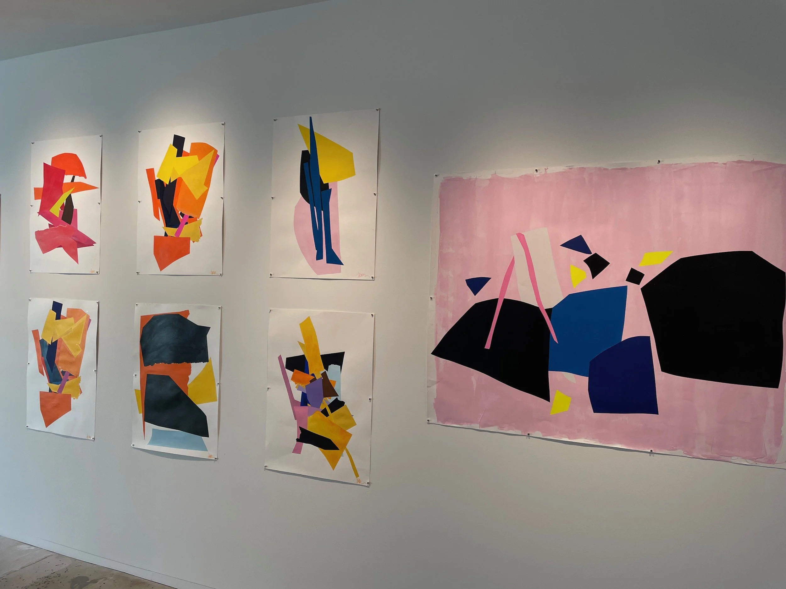 Art gallery wall displaying nine abstract paintings with colorful geometric shapes, alongside a larger pink background piece with black, blue, and pink shapes.