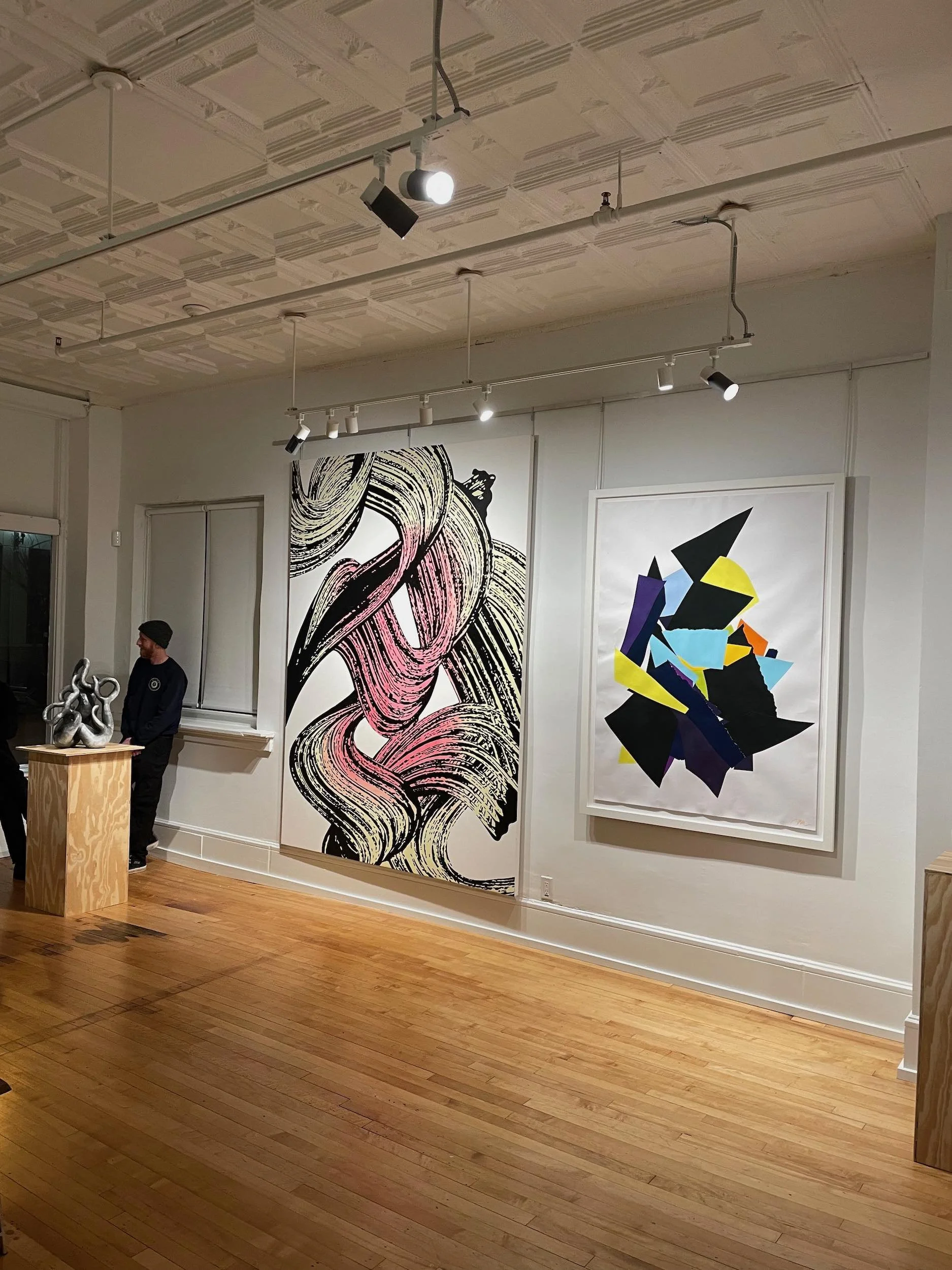 View of an art gallery with hardwood floors and white walls displaying three large abstract artworks. Two people are standing near a pedestal with a shiny sculpture. The ceiling has decorative molding and track lighting illuminating the artworks.