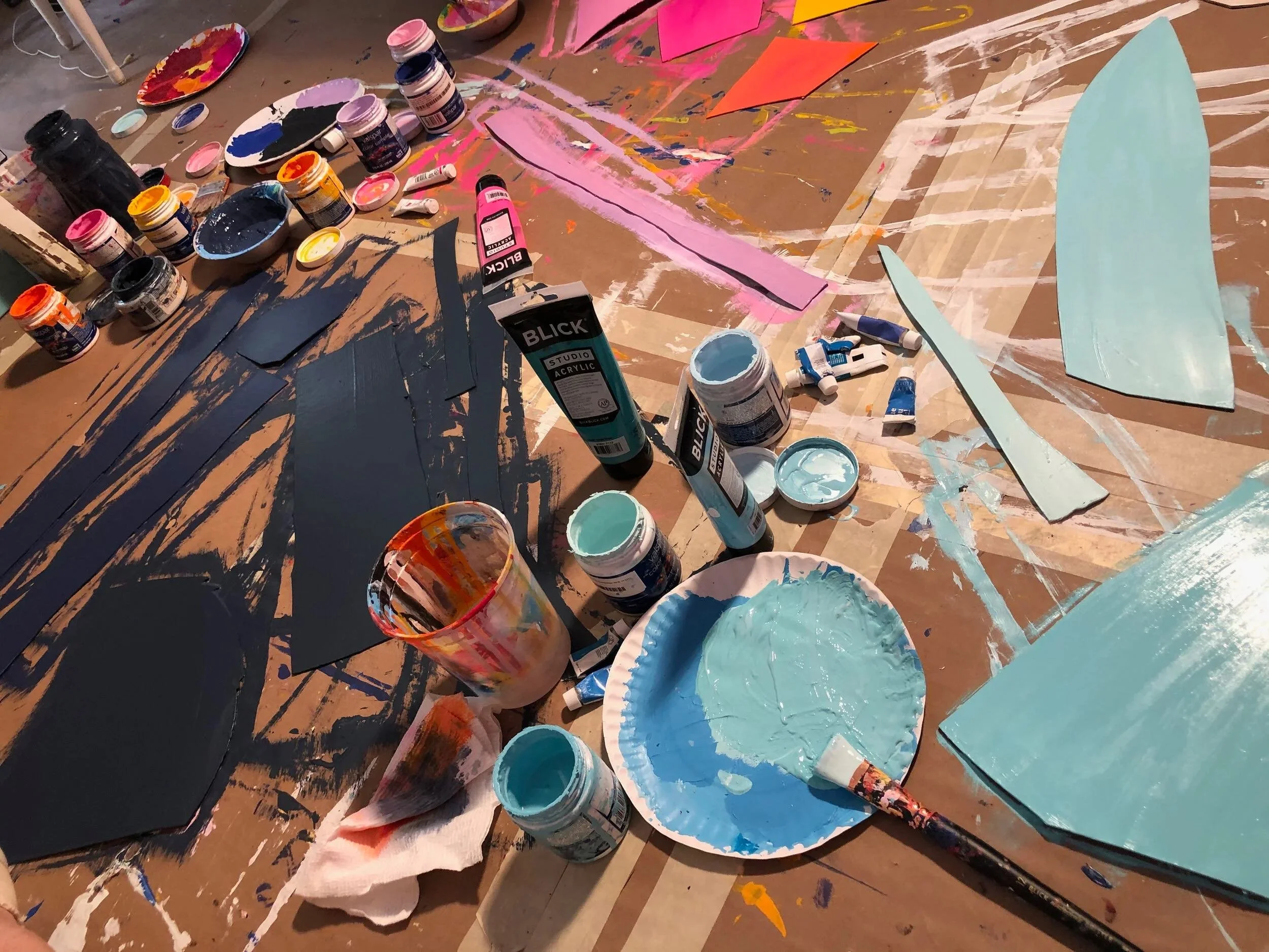 Paint supplies on a table, including jars of acrylic paint, paint tubes, brushes, paper plates with mixed paint, cut paper shapes, and painted foam boards. The table is covered with sketch lines and paint splatters.