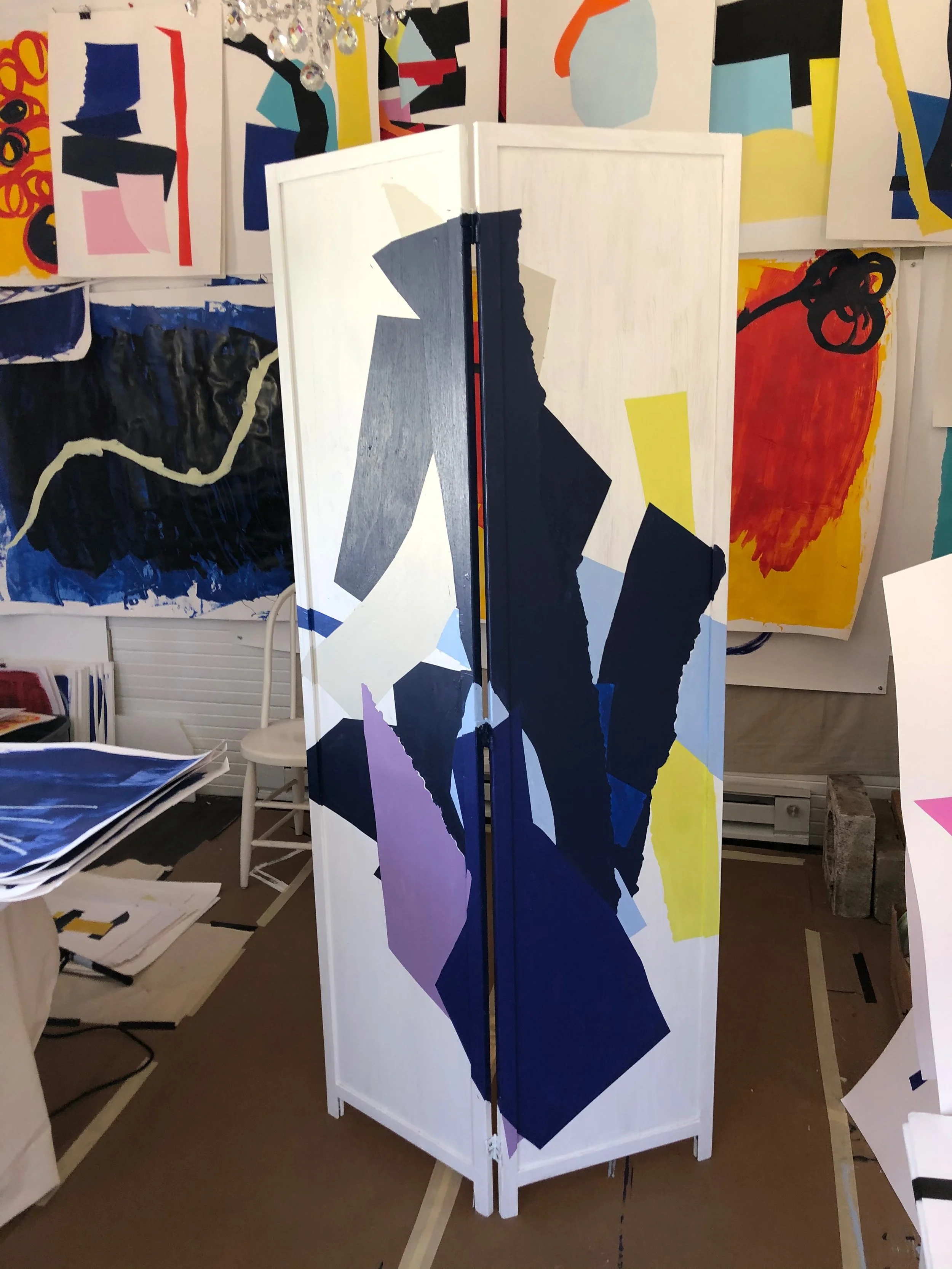 A folding room divider with abstract geometric shapes in shades of blue, purple, black, grey, and yellow painted on it, standing in an art studio with colorful abstract paintings on the walls.