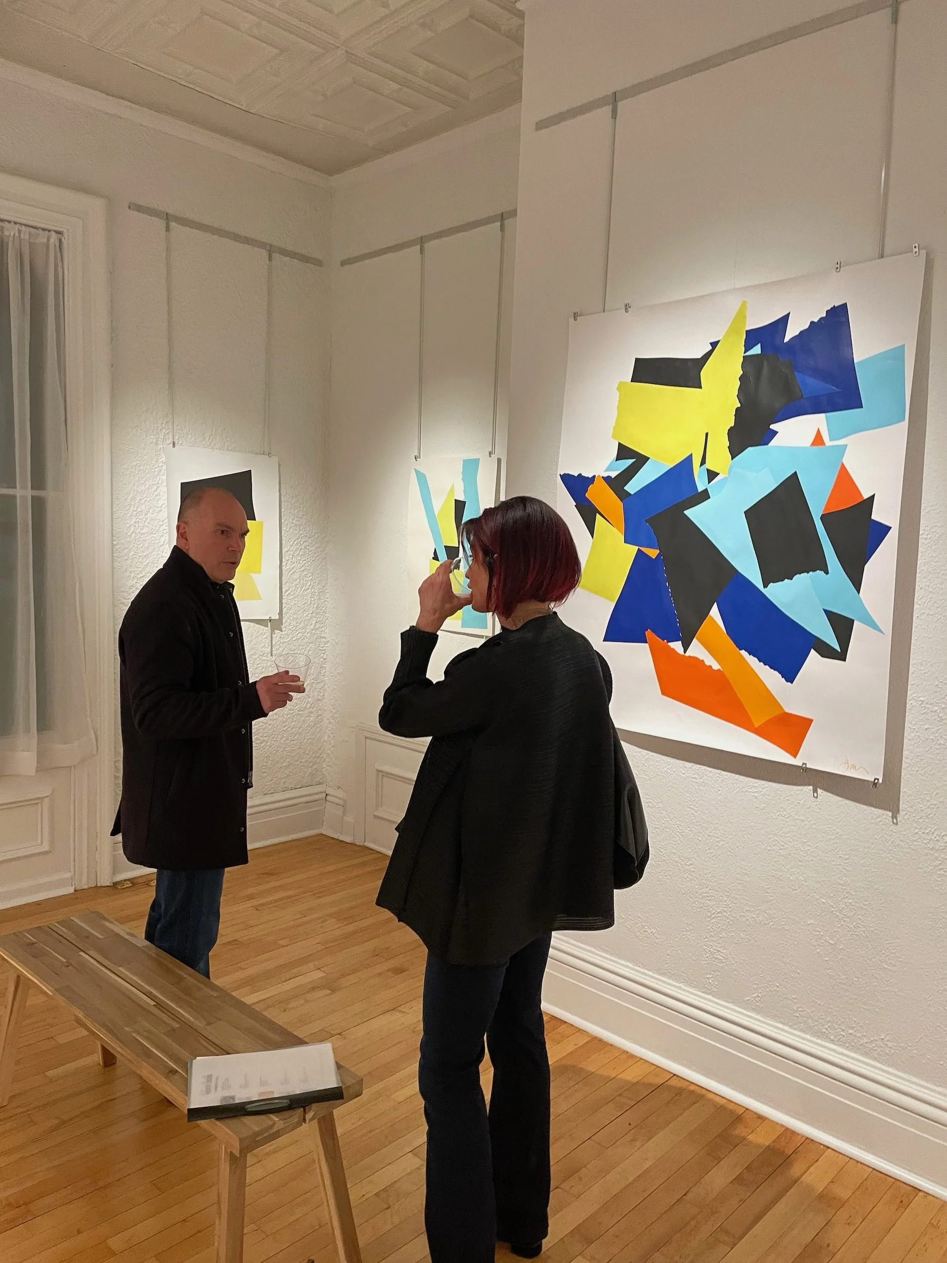 Two people standing and conversing in an art gallery, with colorful abstract paintings on the wall behind them.