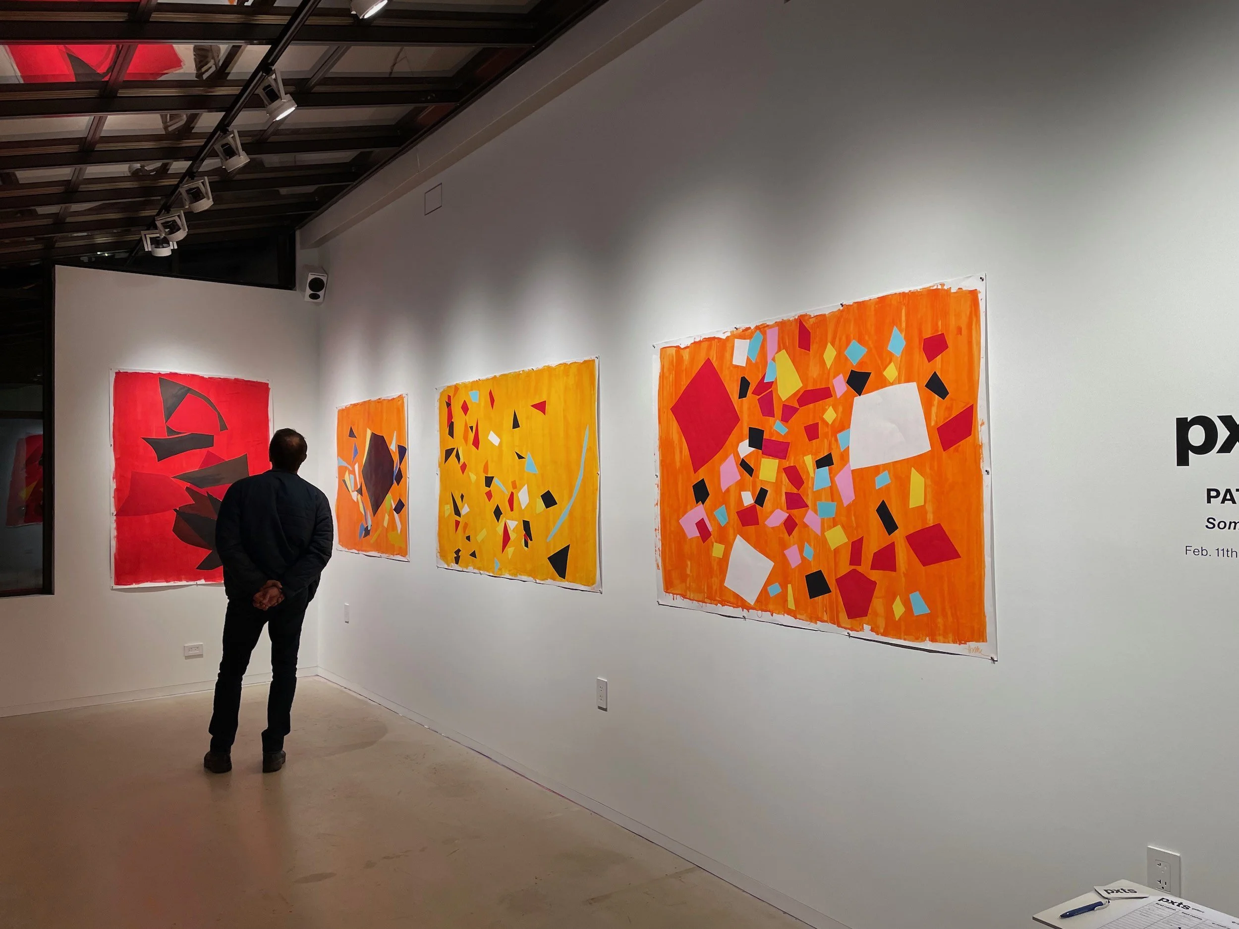 Man in black standing in art gallery observing three colorful abstract paintings on white wall. The paintings feature vibrant colors, geometric shapes, and dynamic compositions.
