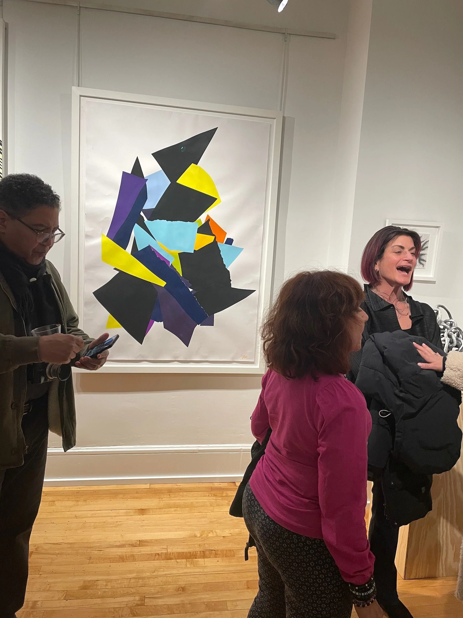 Three people in a gallery space near an abstract colorful artwork on the wall. One woman is smiling and singing, another woman is standing with her back to the camera, and a man is looking at his phone.