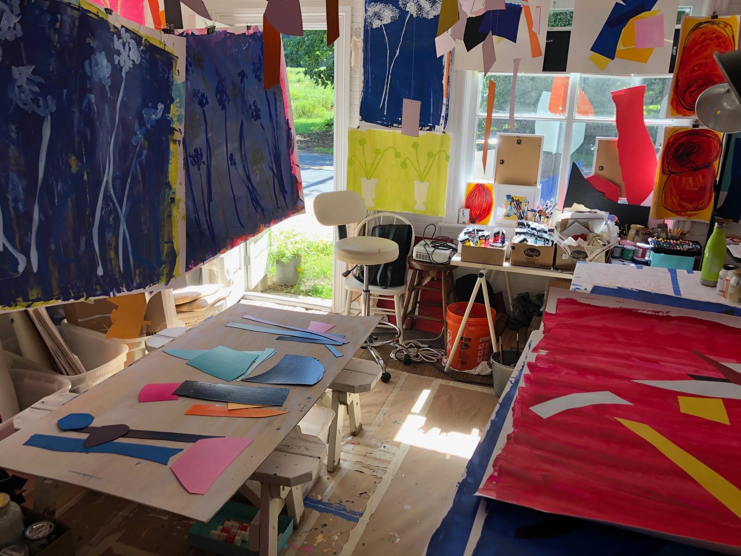 Art studio with colorful paintings of flowers and abstract designs, art supplies scattered around, and sunlight coming through the window.