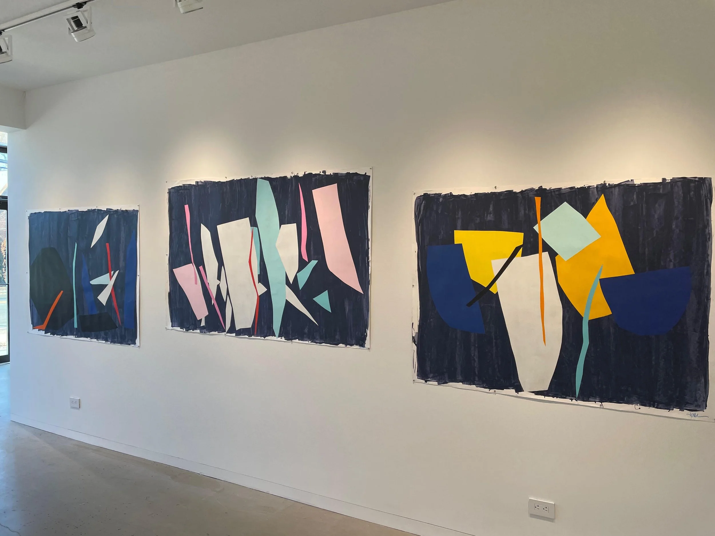 Three large abstract paintings mounted on a white gallery wall. The paintings feature bold geometric shapes in various colors on dark backgrounds.